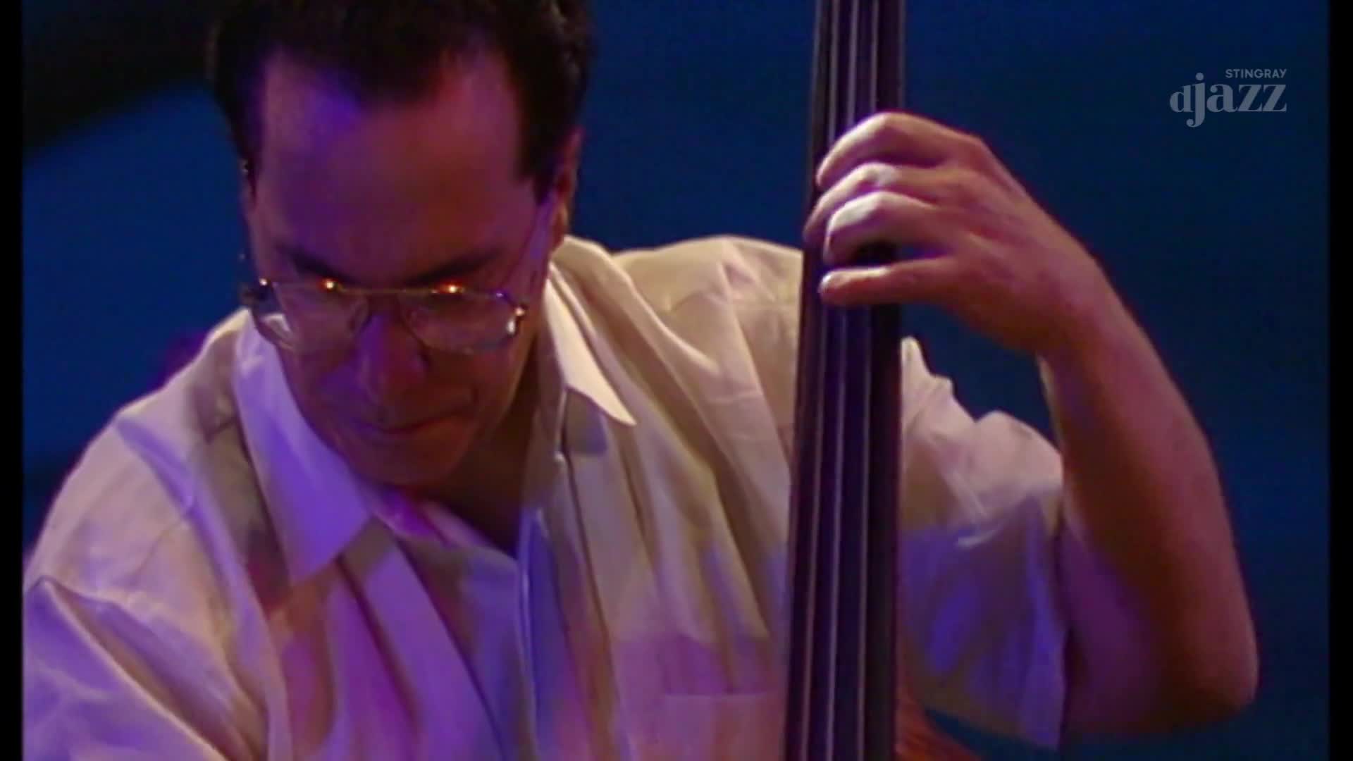 The bassist, in a light-colored shirt, focuses intently on his instrument. His fingers move along the strings of the upright bass as the music from Stingray DJAZZ fills the air.
