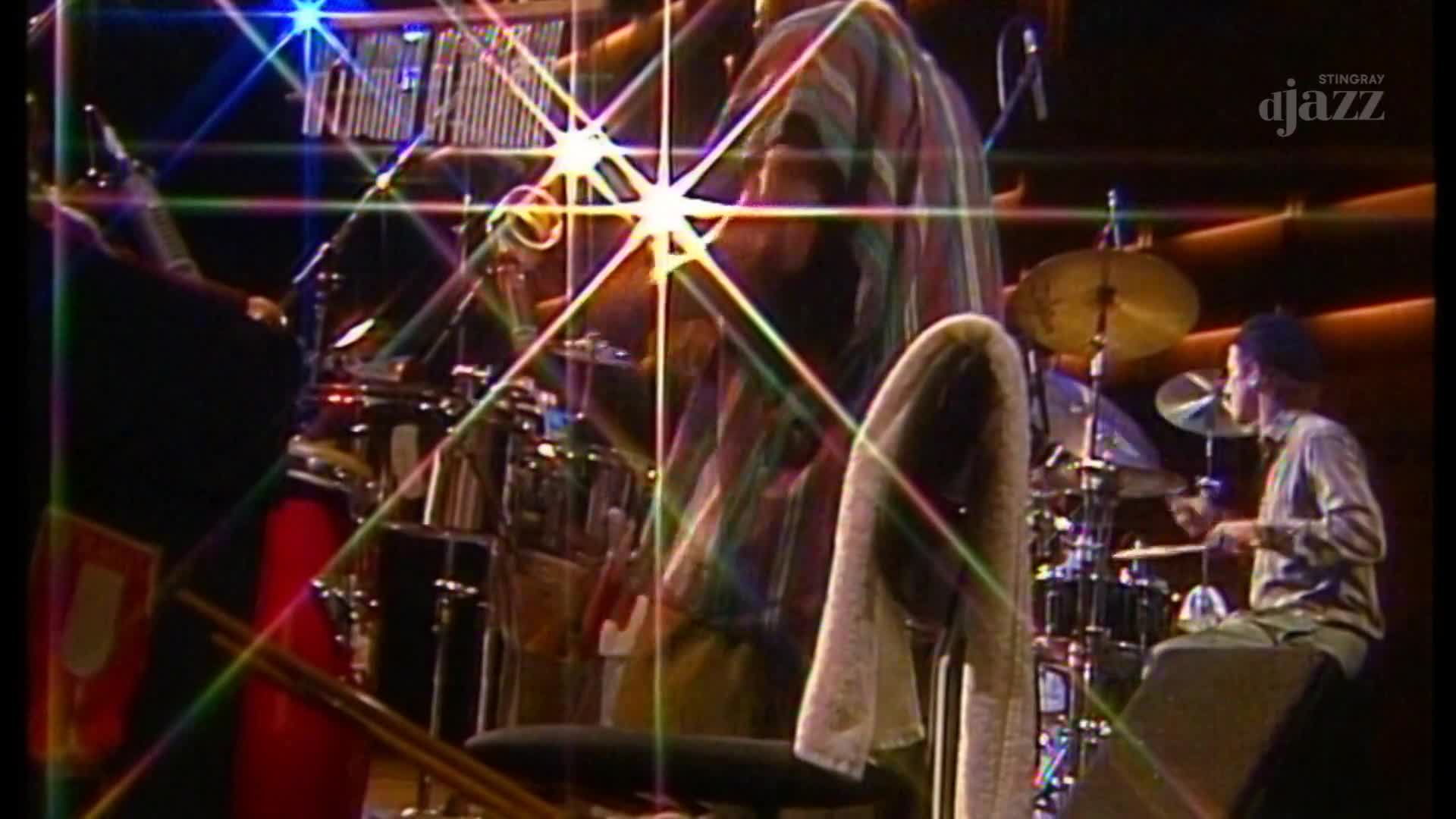 The drummer, lit by stage lights, keeps the rhythm with his sticks, while another musician's back is turned to the audience. A towel hangs over a chair, and the Stingray DJAZZ logo is visible in the corner.
