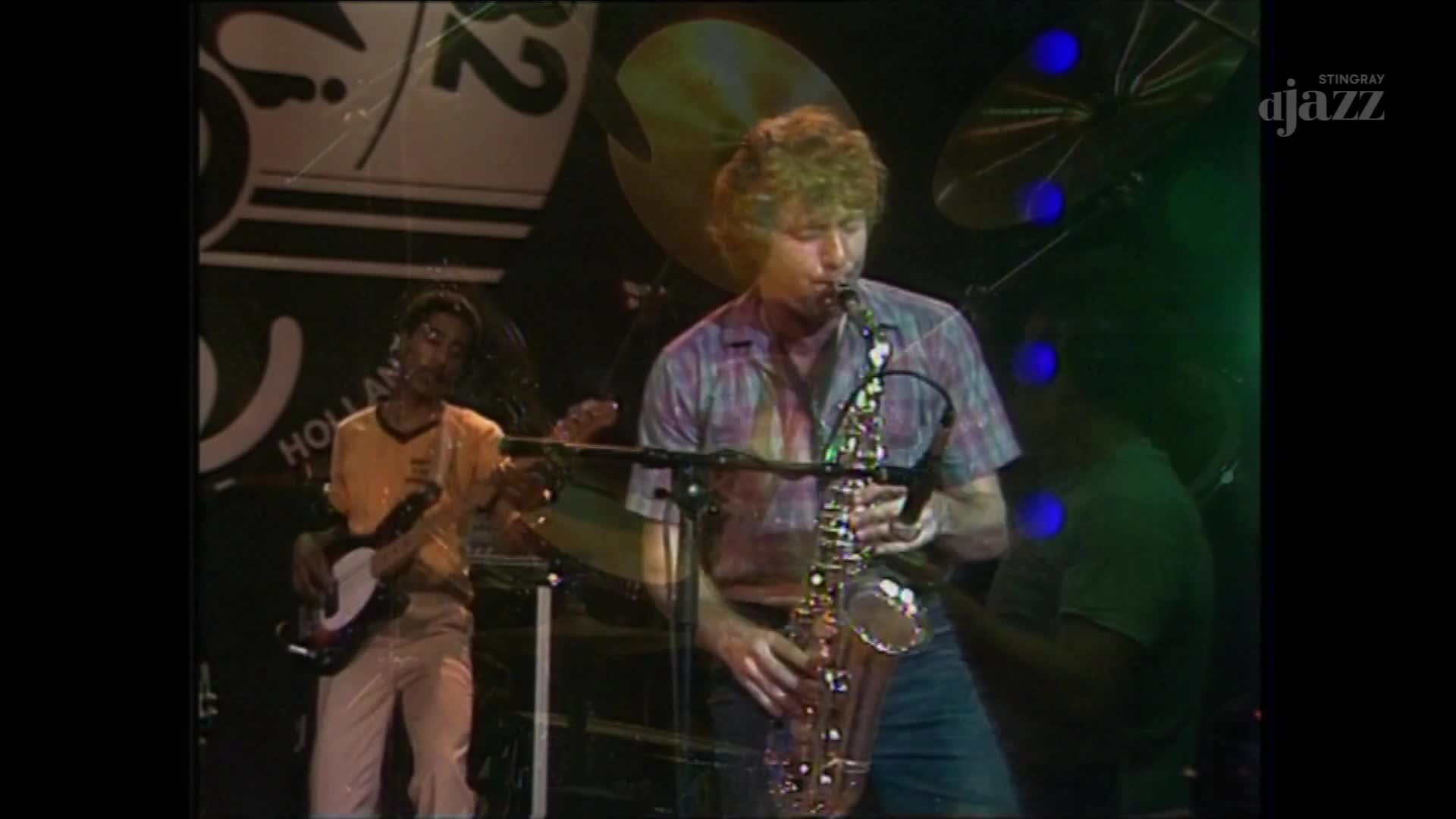 A musician in a plaid shirt blows into a saxophone, his fingers dancing over the keys. Behind him, another player strums a bass guitar, bathed in stage lights.
