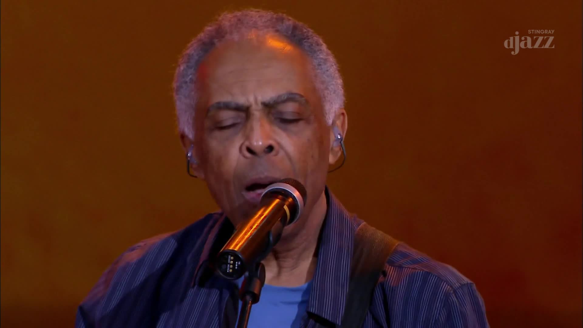 Under the Stingray DJAZZ logo, a musician with closed eyes sings into a microphone. His face is lit, and he's wearing a blue shirt, the strap of a guitar visible over his shoulder.
