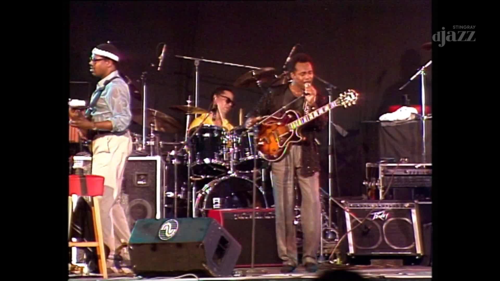 The guitarist in the center is singing into a microphone while strumming his hollow-body electric. Behind him, the drummer in sunglasses keeps the beat, and the other musician stands with his instrument on a stool.
