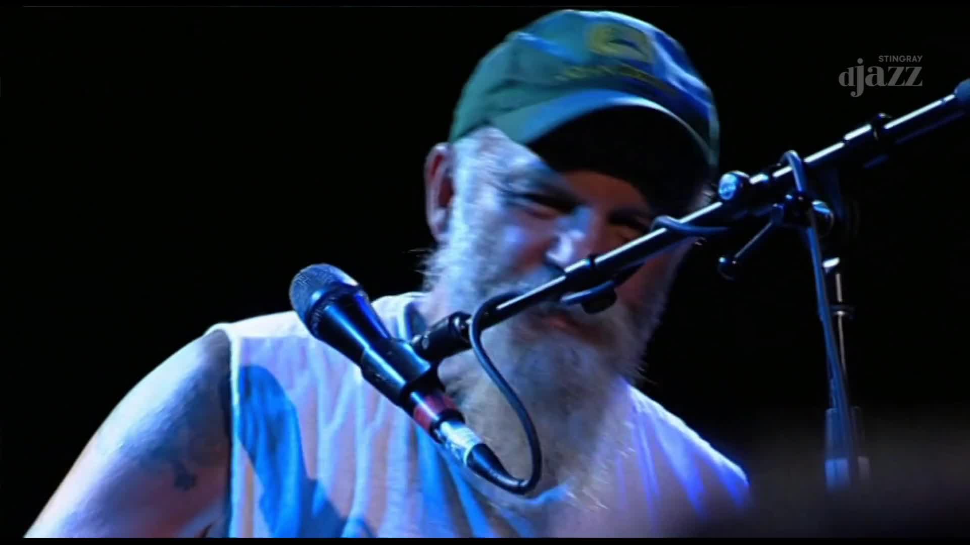 A man in a green cap, beard, and white tank top leans into a microphone, singing. The Stingray DJAZZ logo is visible in the upper right corner, suggesting a Canadian broadcast.
