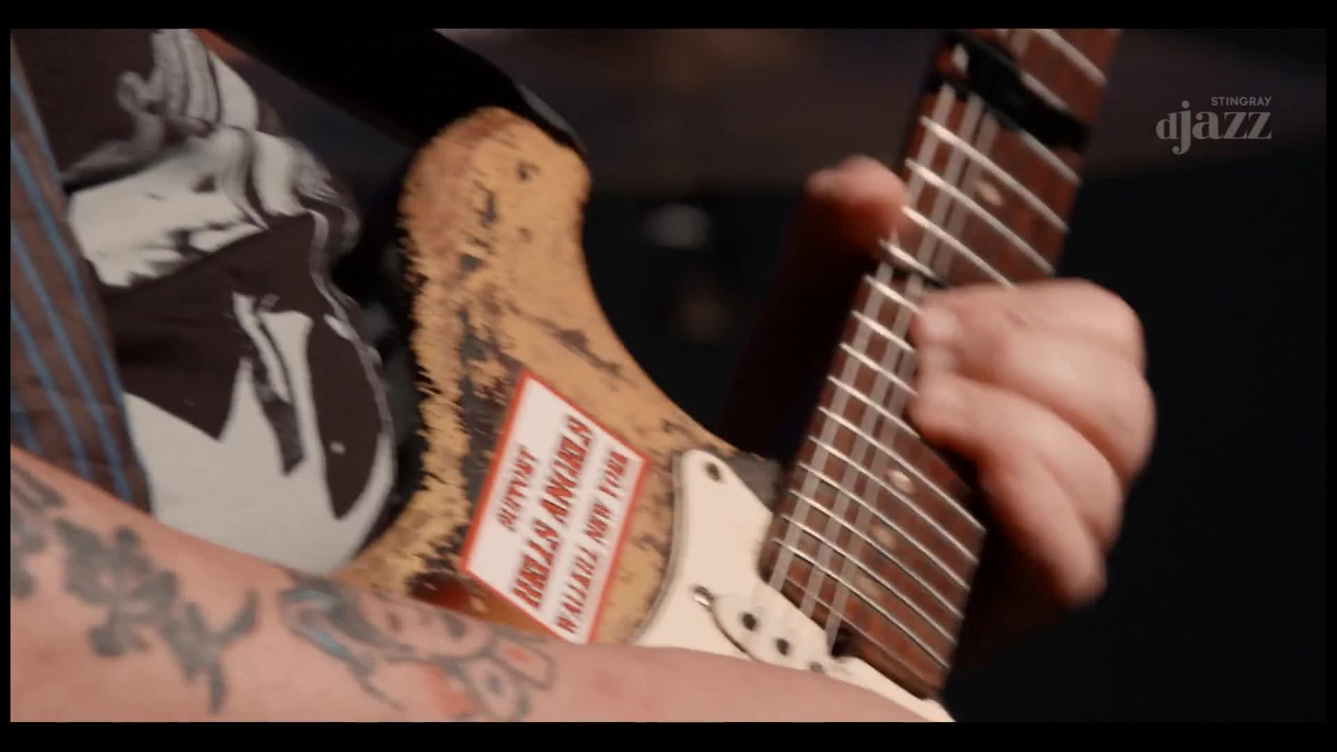A hand, tattooed and weathered, is fretting the neck of a well-worn guitar, the strings vibrating. The Stingray DJAZZ logo is visible as the music unfolds.
