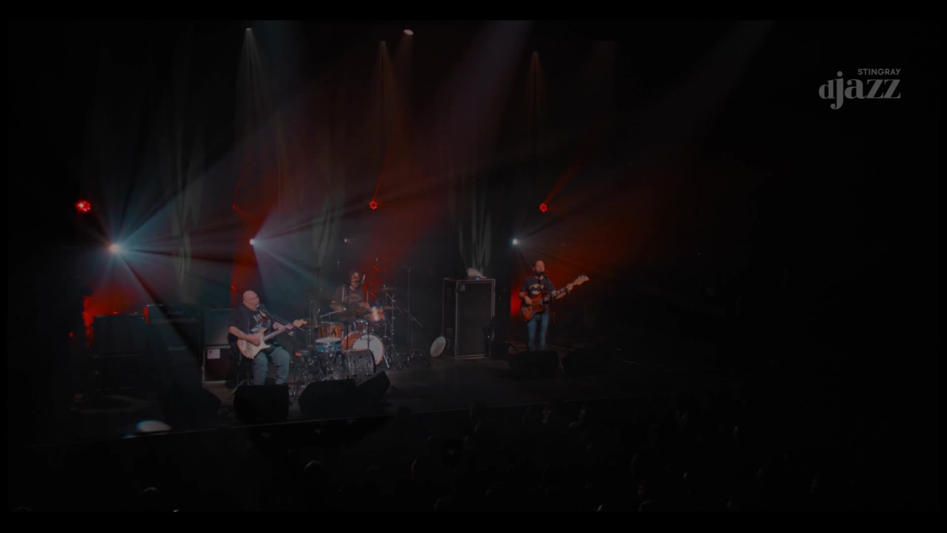 The band, bathed in red and white spotlights, is playing on stage. A bald guitarist strums in front, while the drummer keeps the beat in the back.
