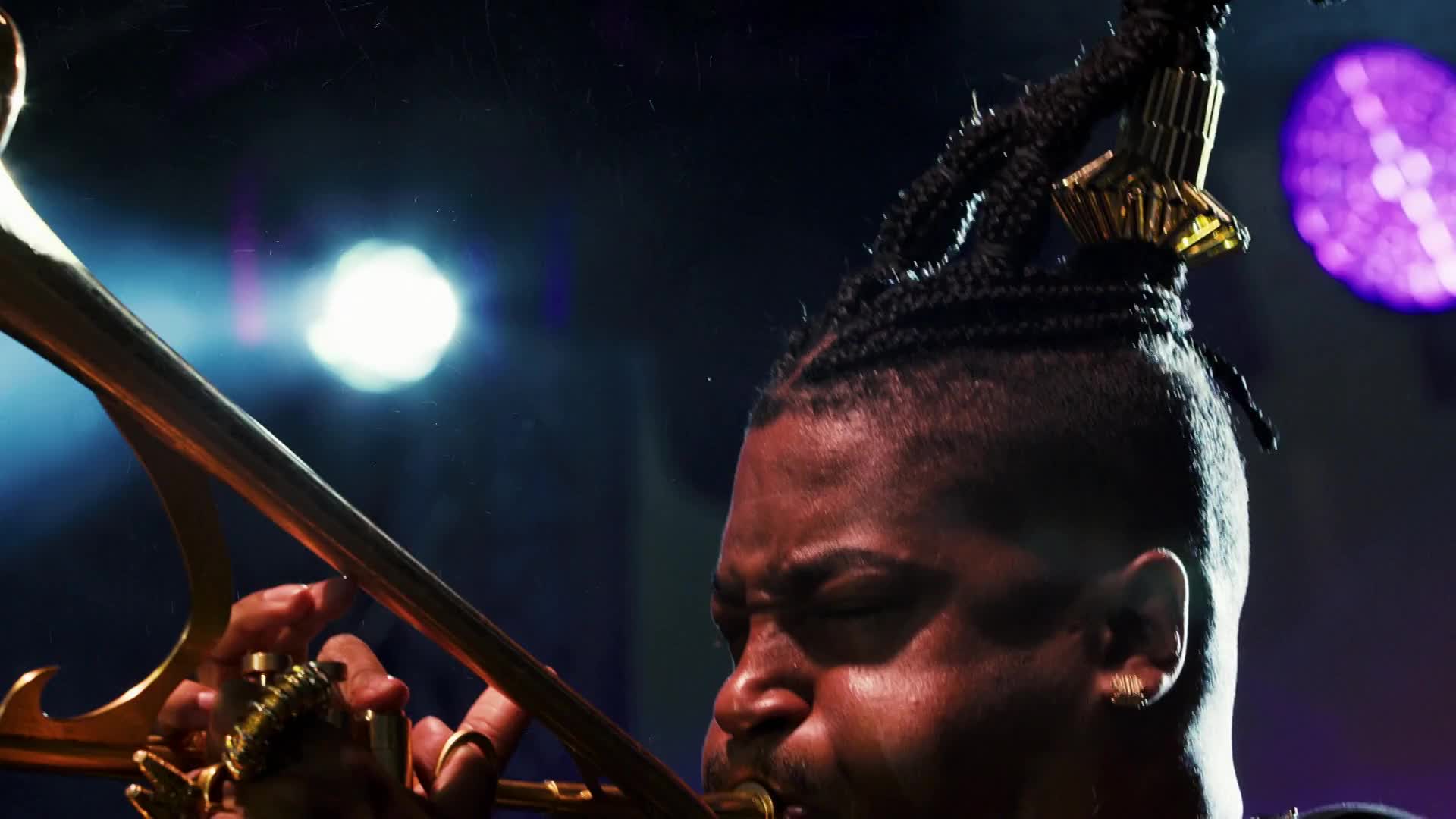The musician's lips are pressed against the mouthpiece, and his fingers work the slide of the trombone. The brass instrument gleams under the stage lights, reflecting the energy of the performance.
