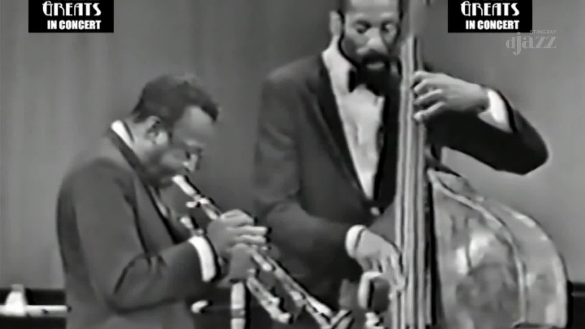 A man in a dark suit blows into a trumpet, his fingers dancing over the valves. Beside him, another man, also in a suit, bows a double bass, the instrument's curves gleaming under the stage lights.
