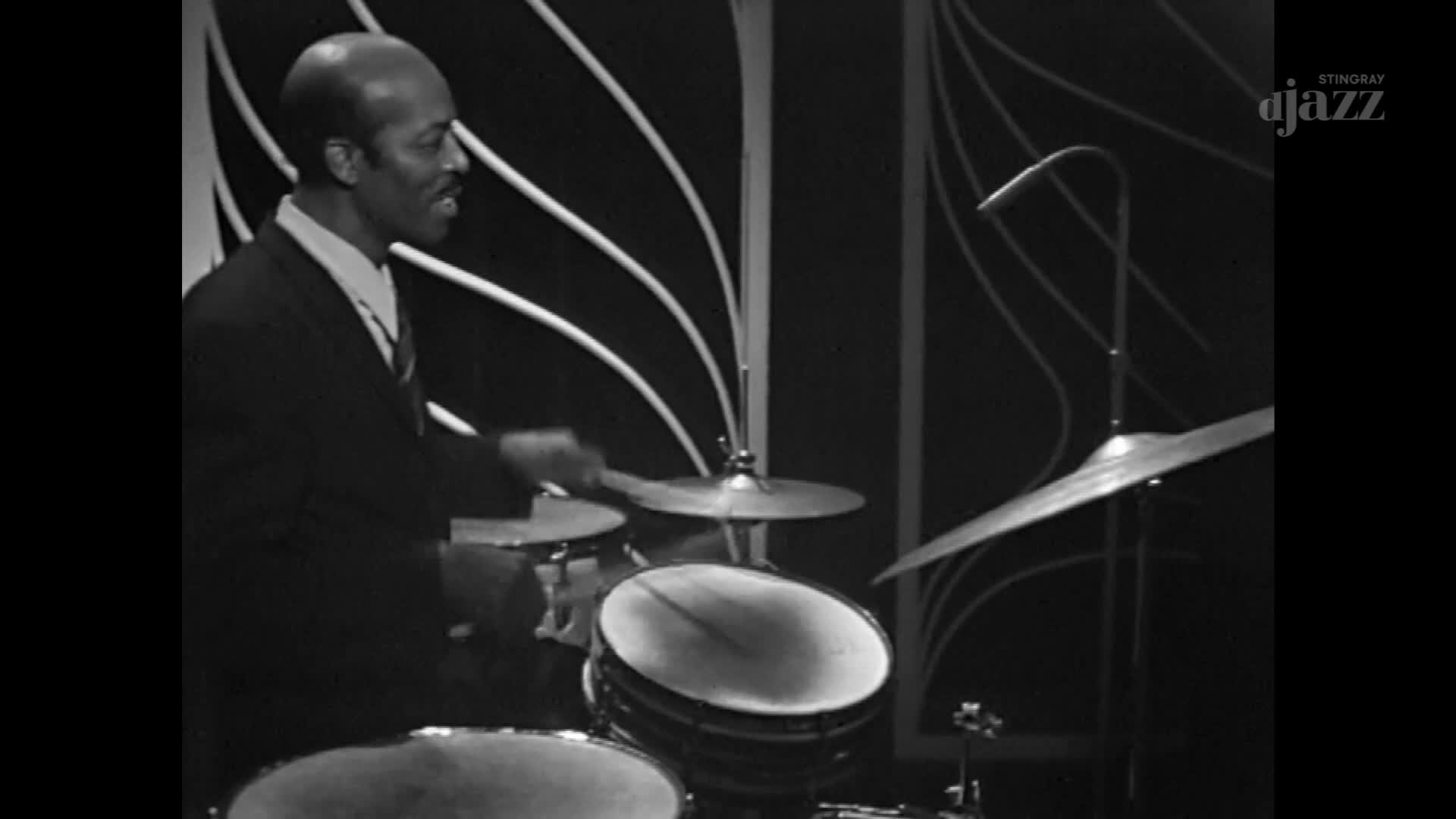A drummer in a suit is focused, his sticks striking the drums and cymbals. The Stingray DJAZZ logo is visible in the corner.
A drummer in a suit is focused, his sticks striking the drums and cymbals. The Stingray DJAZZ logo is visible in the corner.