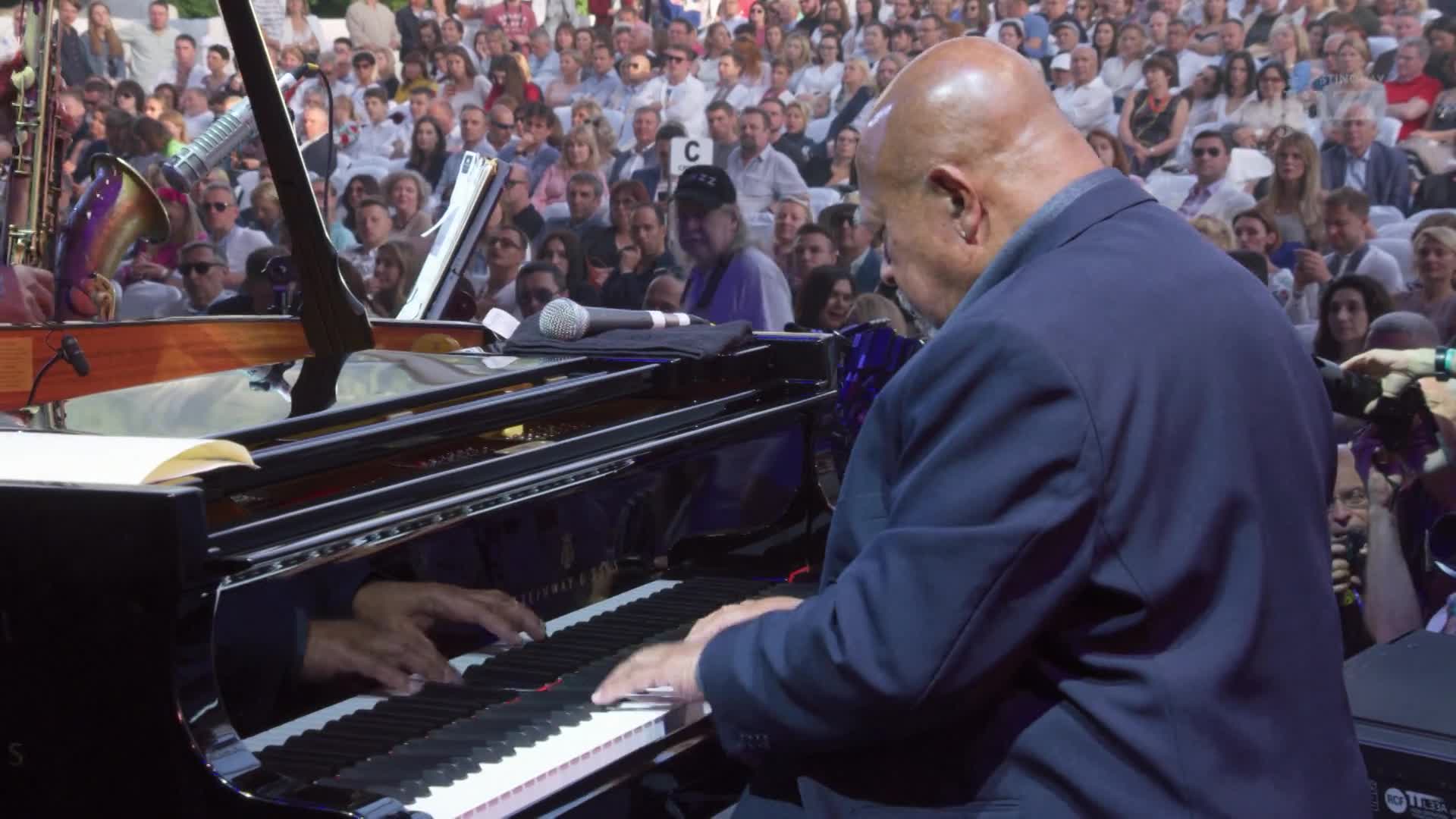 A man in a blue suit is hunched over a grand piano, his fingers dancing across the keys. The audience, packed tightly in the background, watches intently as the music fills the outdoor venue in Canada, captured by Stingray DJAZZ.
A man in a blue suit is hunched over a grand piano, his fingers dancing across the keys. The audience, packed tightly in the background, watches intently as the music fills the outdoor venue in Canada, captured by Stingray DJAZZ.