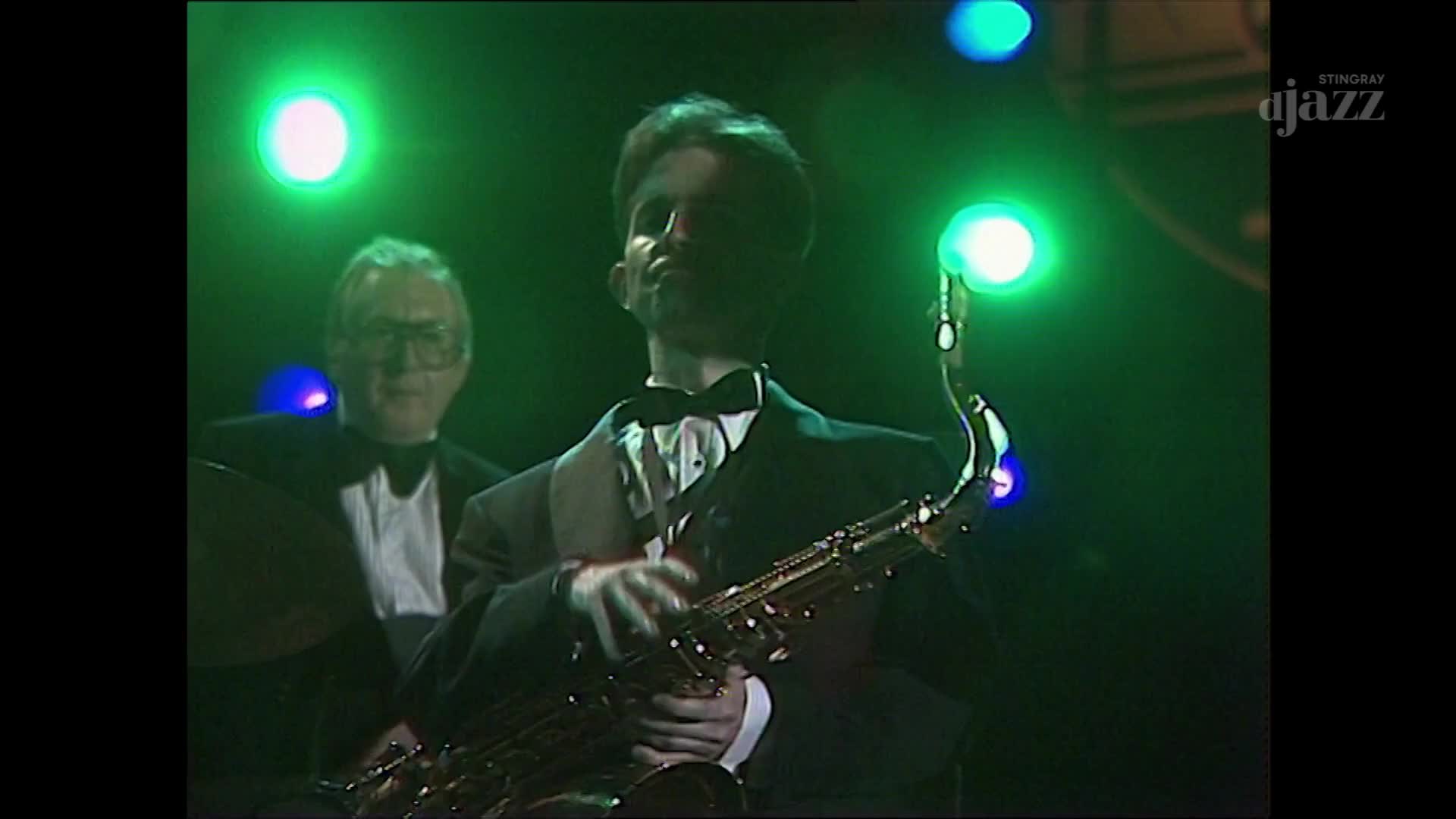 The saxophonist in a tuxedo holds his instrument, fingers dancing over the keys as he plays. Behind him, a drummer in a similar outfit keeps the beat, illuminated by the green stage lights.
The saxophonist in a tuxedo holds his instrument, fingers dancing over the keys as he plays. Behind him, a drummer in a similar outfit keeps the beat, illuminated by the green stage lights.