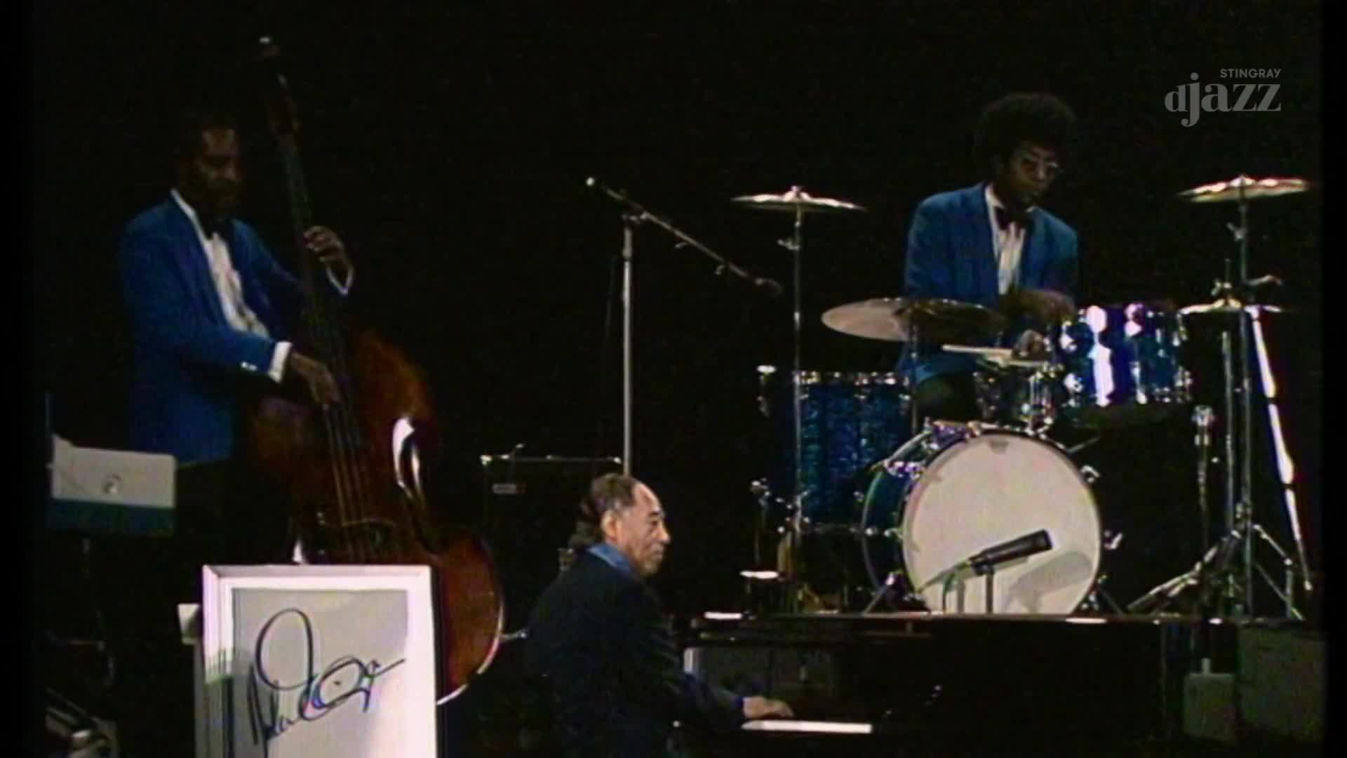 The bassist, in a bright blue jacket, draws his bow across the strings of the upright bass. The drummer's sticks tap a steady rhythm on the blue-sparkle drums, while the pianist's fingers dance across the keys.
The bassist, in a bright blue jacket, draws his bow across the strings of the upright bass. The drummer's sticks tap a steady rhythm on the blue-sparkle drums, while the pianist's fingers dance across the keys.