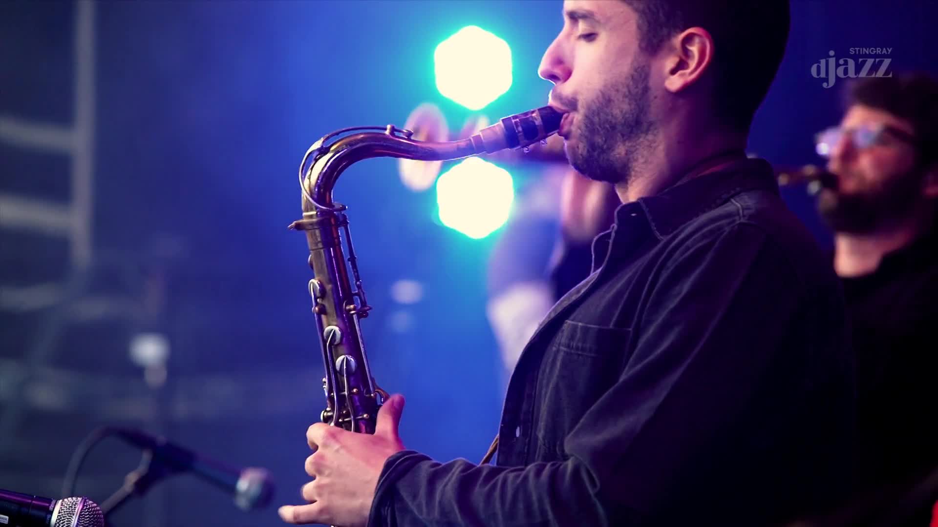 The saxophonist is blowing into his instrument, his breath visible in the stage lights. Behind him, another musician plays, part of the band performing on Stingray DJAZZ from Canada.
