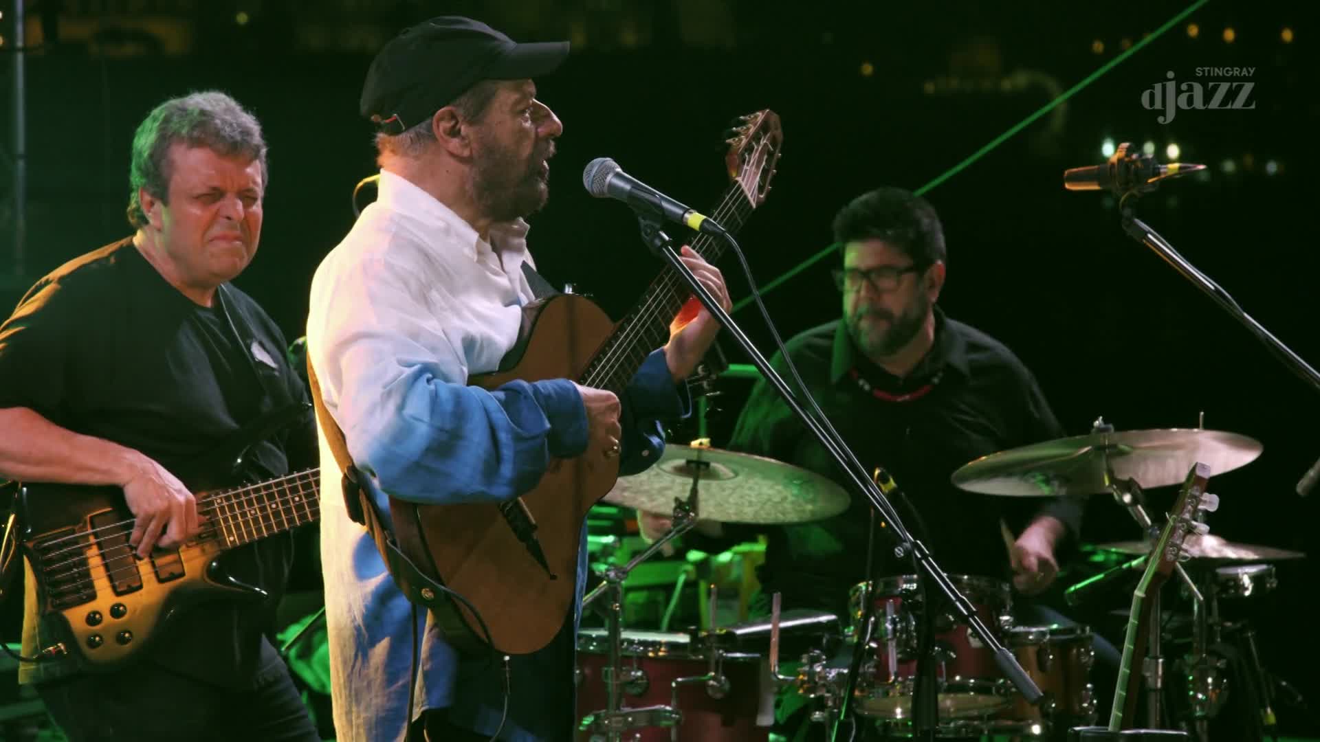 The guitarist in the center is singing into a microphone, his hands moving over the fretboard of his acoustic guitar. The drummer's sticks are a blur as they hit the cymbals and drums, keeping the beat for the live performance from Canada, broadcast on Stingray DJAZZ.
