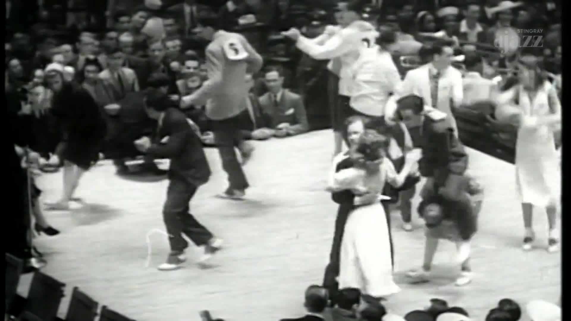 Couples are dancing on a large, open floor, some in close embraces, others with more energetic steps. A man in a suit is bouncing on his feet, while another with a number on his back moves quickly across the floor.
