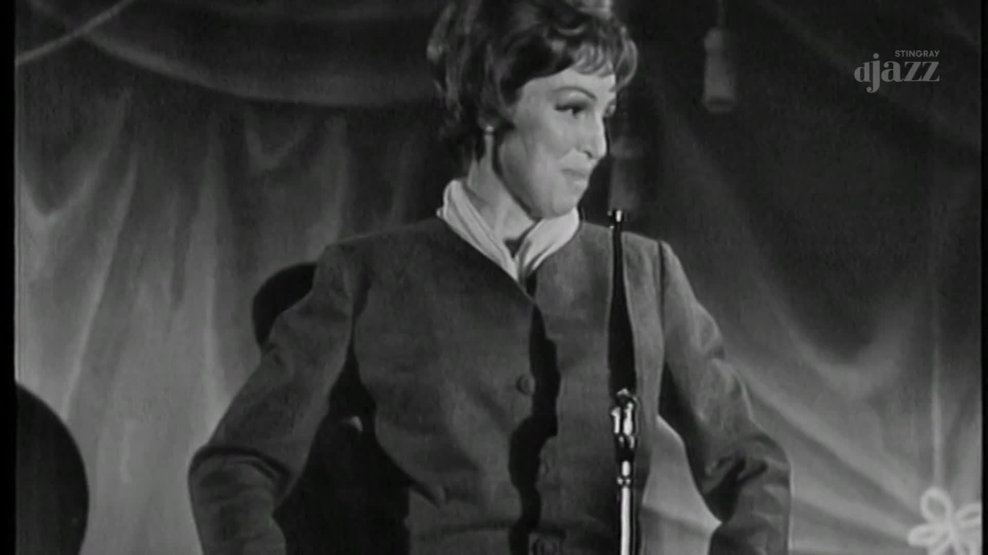 A woman in a blazer stands before a microphone, looking to her right with a slight smile. The Stingray DJAZZ logo is visible in the corner, suggesting a Canadian jazz performance.
