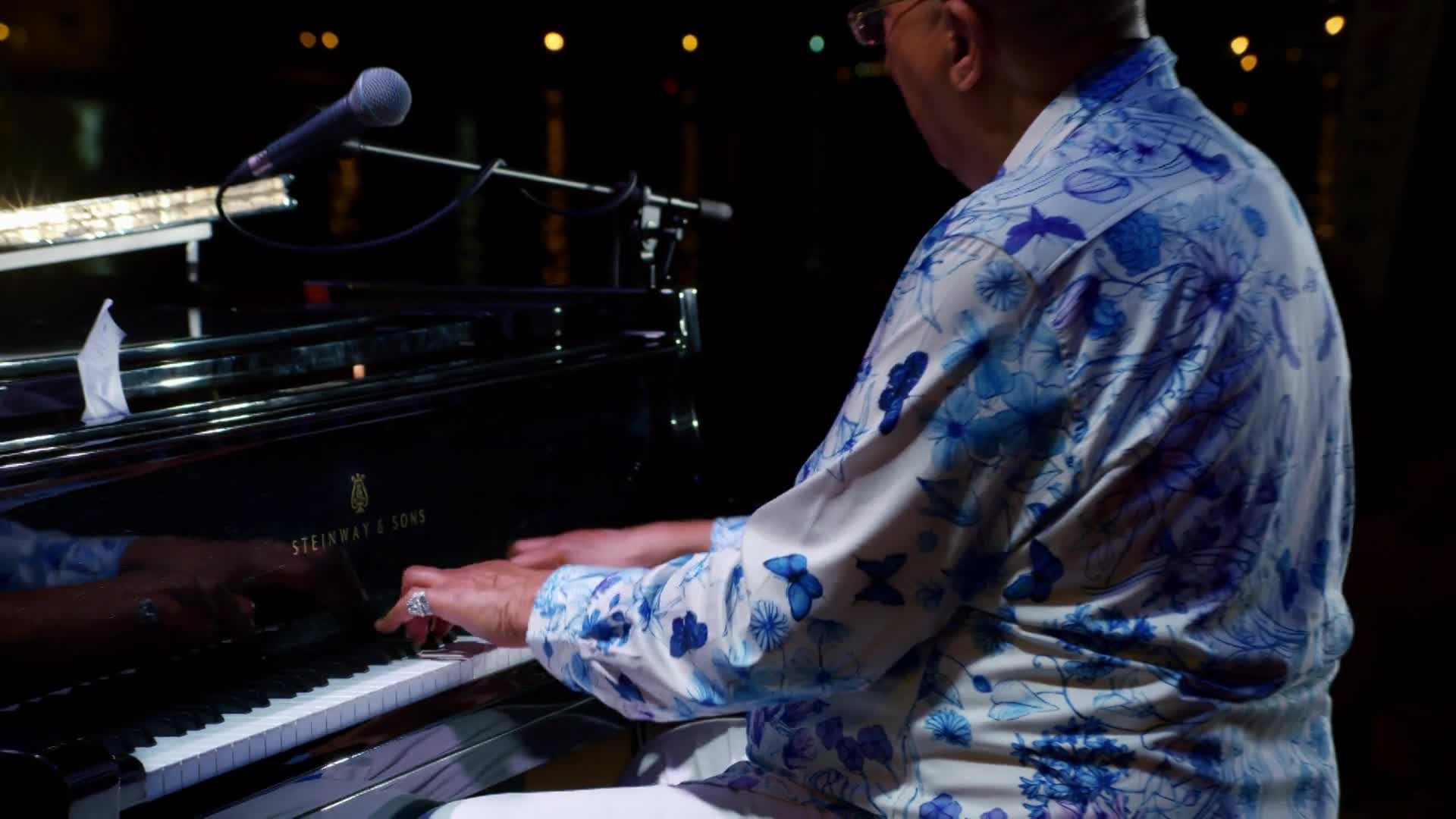 The musician's hands dance across the piano keys, the notes echoing in the dim space. His patterned shirt, a splash of blue and white, contrasts with the black Steinway.
