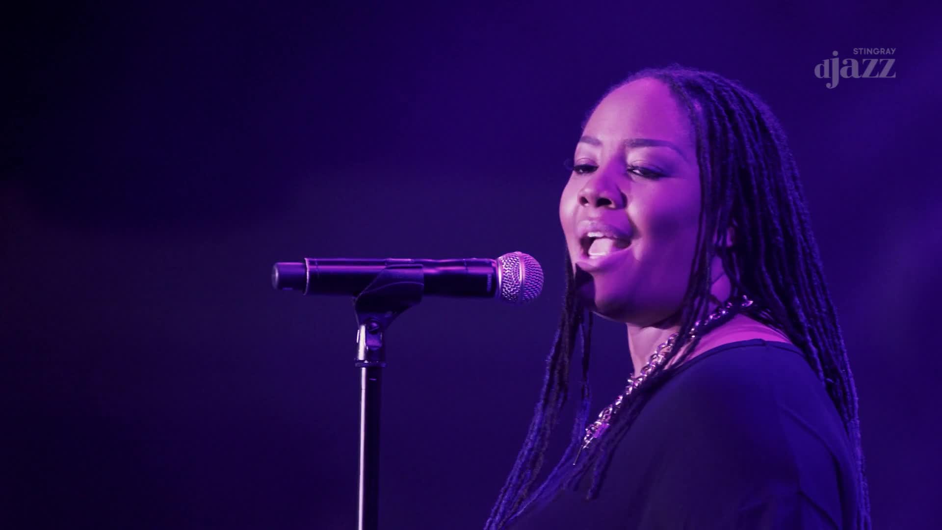 A woman with long braids sings into a microphone, her mouth open in mid-note. The Stingray DJAZZ logo is visible in the corner, and the stage is bathed in purple light.
