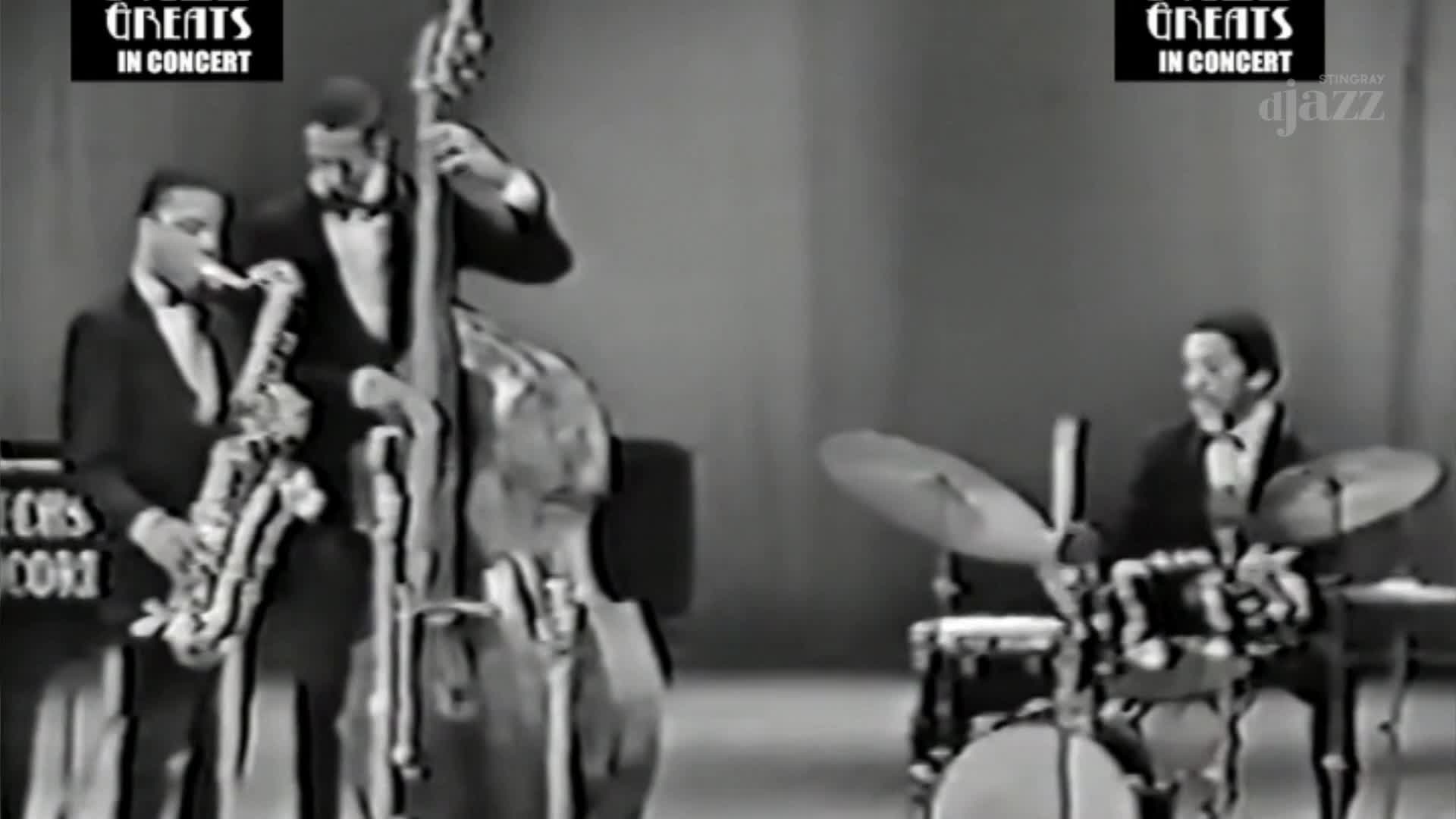 A saxophone player and a bassist stand shoulder-to-shoulder, while a drummer keeps time on the far right. The band, part of a Canadian broadcast on Stingray DJAZZ, is dressed in matching suits.
