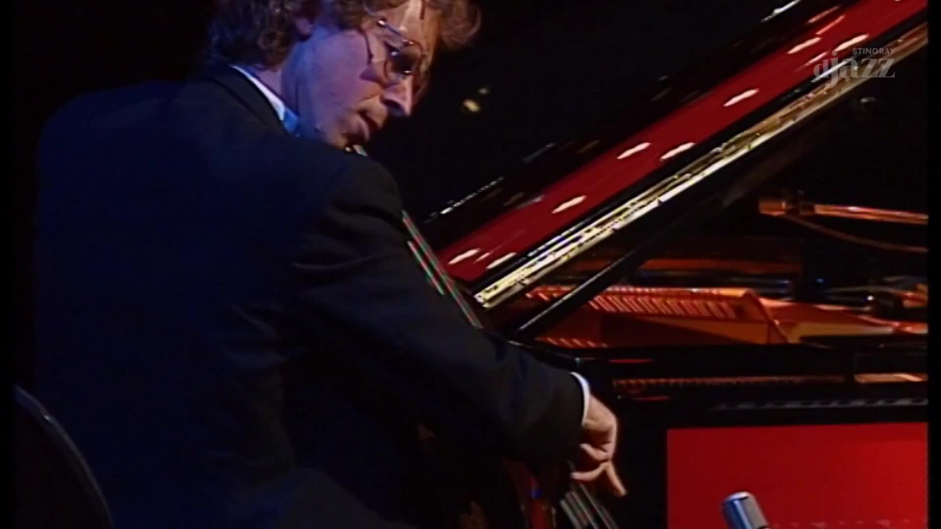 A man in a suit, lit by stage lights, plays a grand piano. His fingers dance across the keys, the polished red surface reflecting the glow of the Stingray DJAZZ set.
