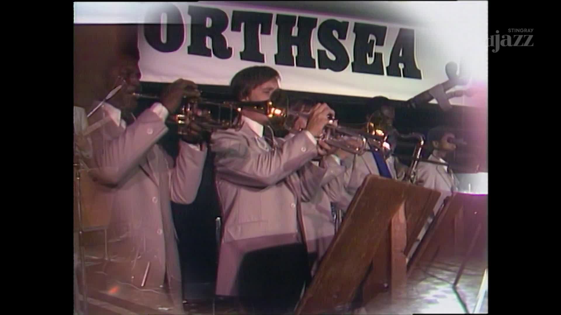 A band in matching jackets is mid-song, trumpets raised and glinting under the stage lights. The "NORTHSEA" banner hangs above them, and the Stingray DJAZZ logo is visible in the corner.
A band in matching jackets is mid-song, trumpets raised and glinting under the stage lights. The "NORTHSEA" banner hangs above them, and the Stingray DJAZZ logo is visible in the corner.