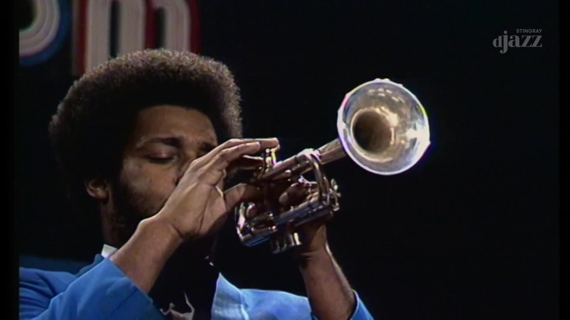 A trumpet player with a large afro, dressed in a bright blue jacket, brings his instrument to his lips. The polished brass gleams under the stage lights as he prepares to play.
