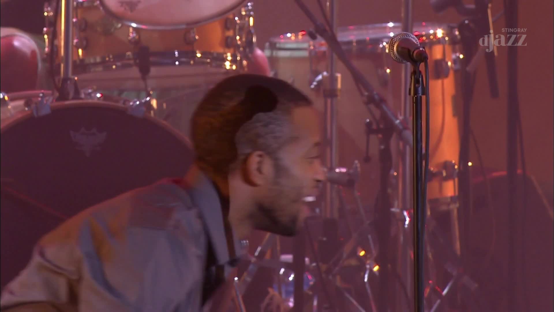 A man leans into a microphone, his face illuminated by stage lights. Behind him, a drum kit gleams under the warm glow, ready for the next beat of the Stingray DJAZZ performance. A man leans into a microphone, his face illuminated by stage lights. Behind him, a drum kit gleams under the warm glow, ready for the next beat of the Stingray DJAZZ performance.