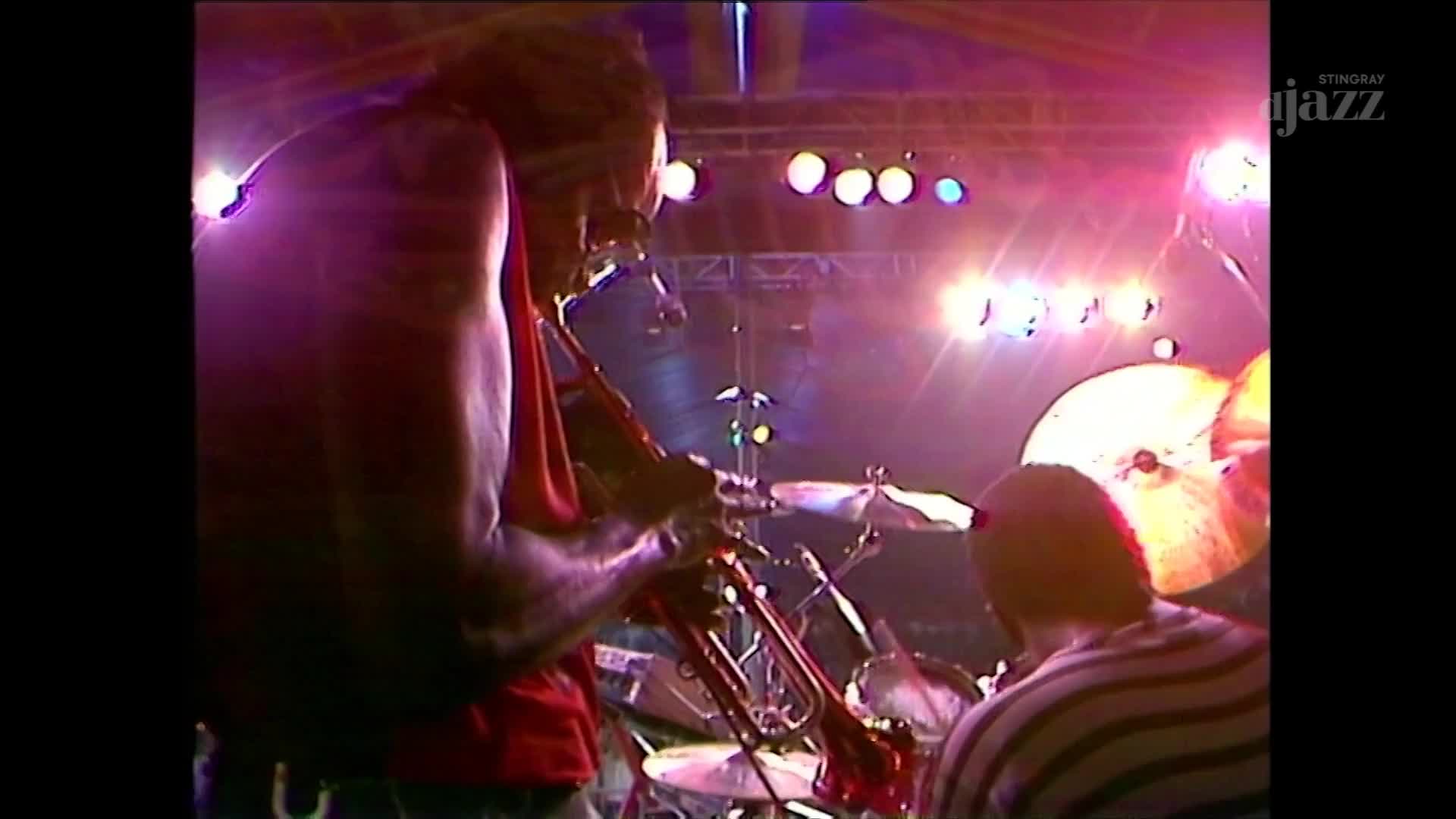 A trumpeter in a red tank top plays his instrument under bright stage lights. Behind him, a drummer's cymbals catch the light as the Stingray DJAZZ broadcast continues. A trumpeter in a red tank top plays his instrument under bright stage lights. Behind him, a drummer's cymbals catch the light as the Stingray DJAZZ broadcast continues.