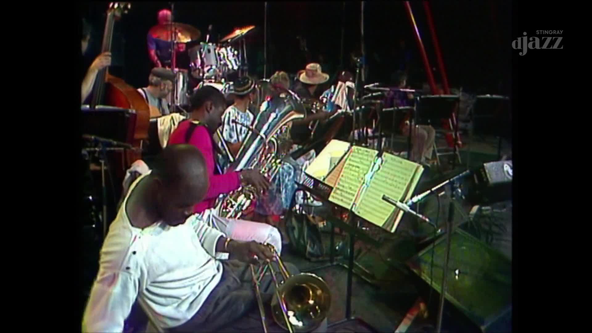 A trombonist in a white shirt leans forward, his instrument poised. Behind him, a tuba player in a bright pink vest is about to play.
