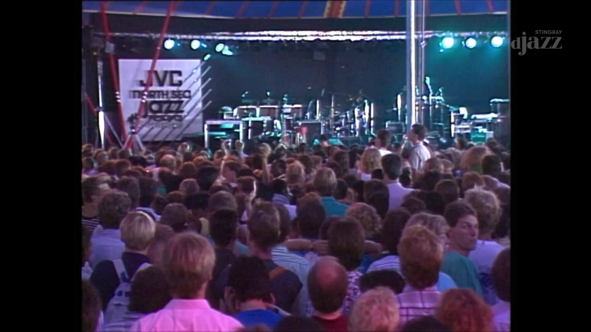 A large crowd gathers under a striped canopy, their faces turned towards a stage set with instruments. The JVC North Sea Jazz Festival logo is prominent on a banner behind the band's equipment.