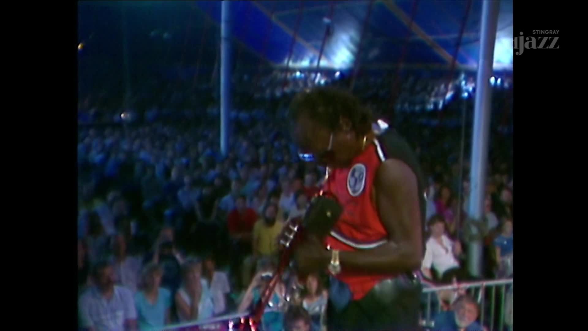 A musician in a red vest tunes his instrument under a tent at a Canadian festival. A large crowd watches from behind a railing.