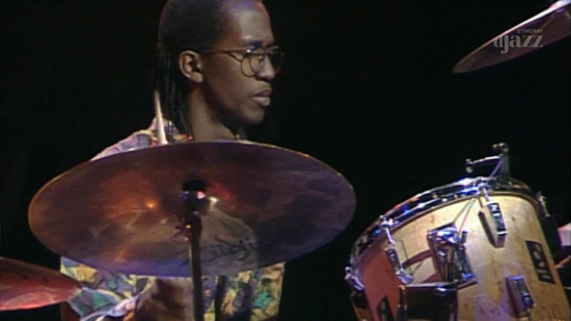 A drummer, his glasses reflecting the stage lights, plays a steady beat on his kit. The Stingray DJAZZ logo appears in the upper right corner.