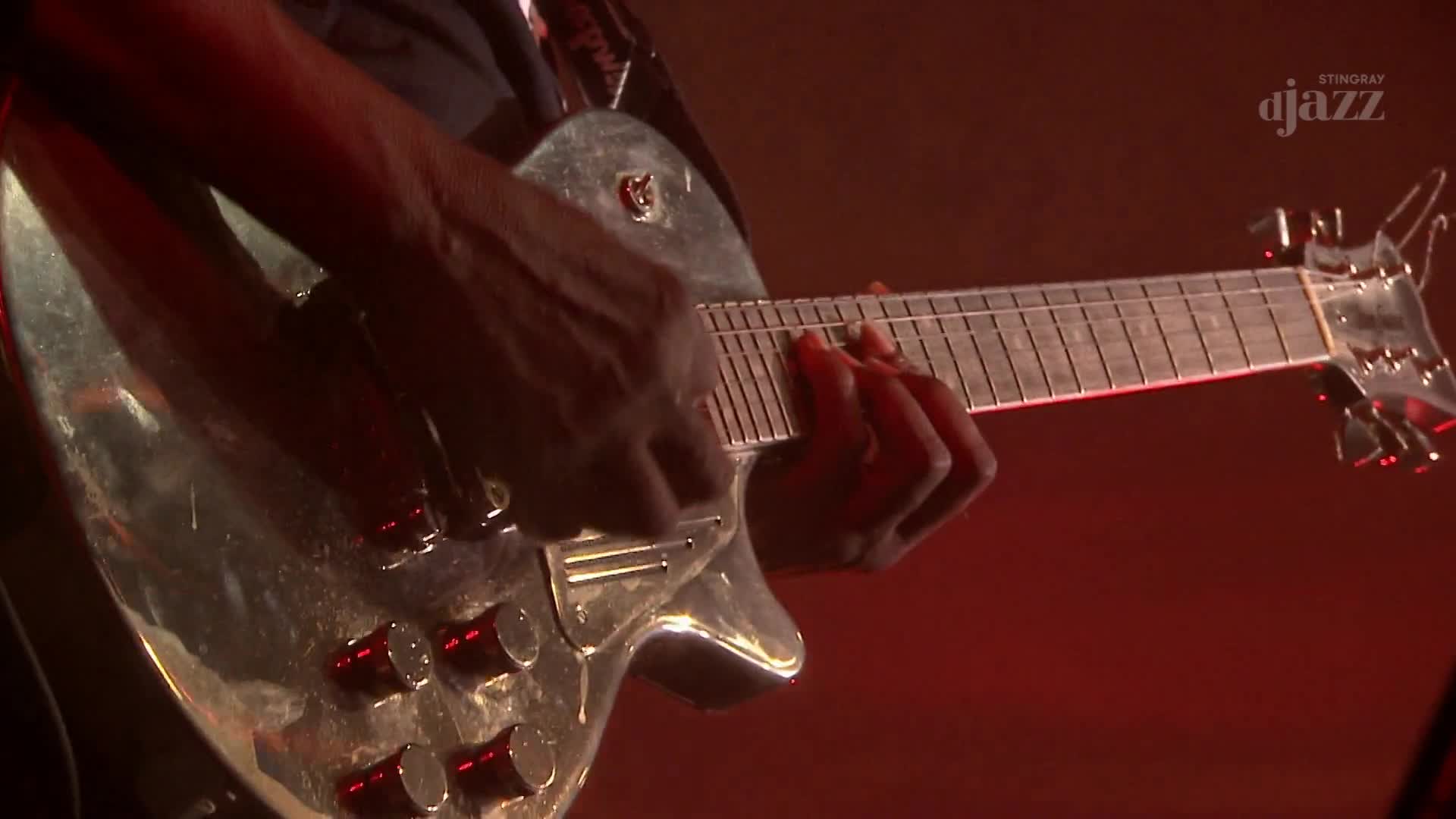 Fingers dance across the fretboard of a gleaming silver guitar. The red stage lights catch the polished surface as the musician plays.