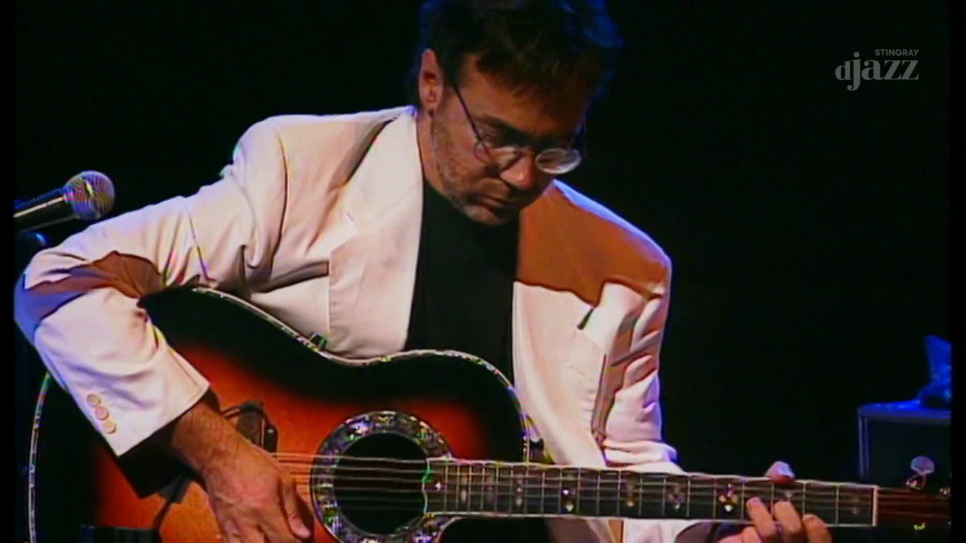 A man in a white jacket plays an acoustic guitar, his fingers moving across the fretboard. The Stingray DJAZZ logo appears in the corner, suggesting a performance broadcast from Canada.
