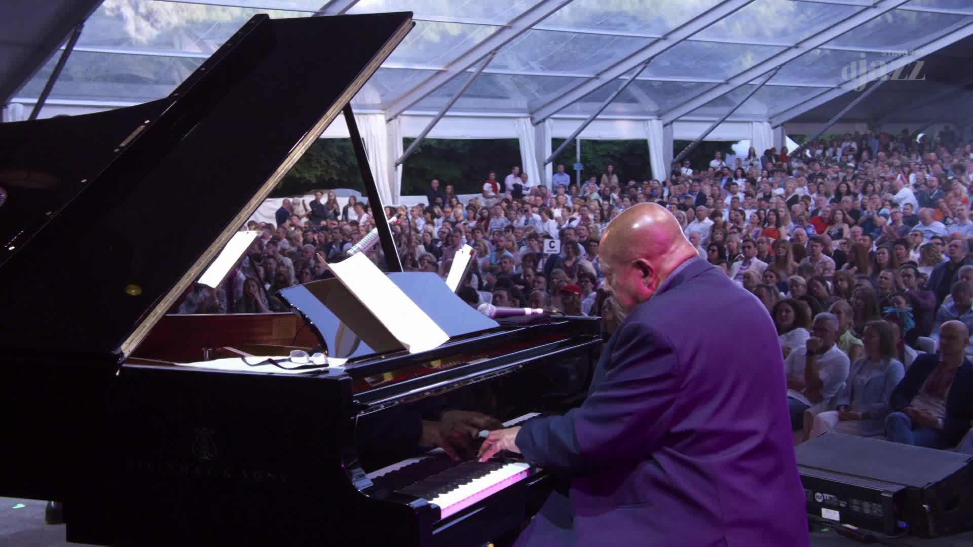 A bald man in a purple suit plays a grand piano under a tent in Canada, a large audience watching. The Stingray DJAZZ logo is visible on a distant speaker.