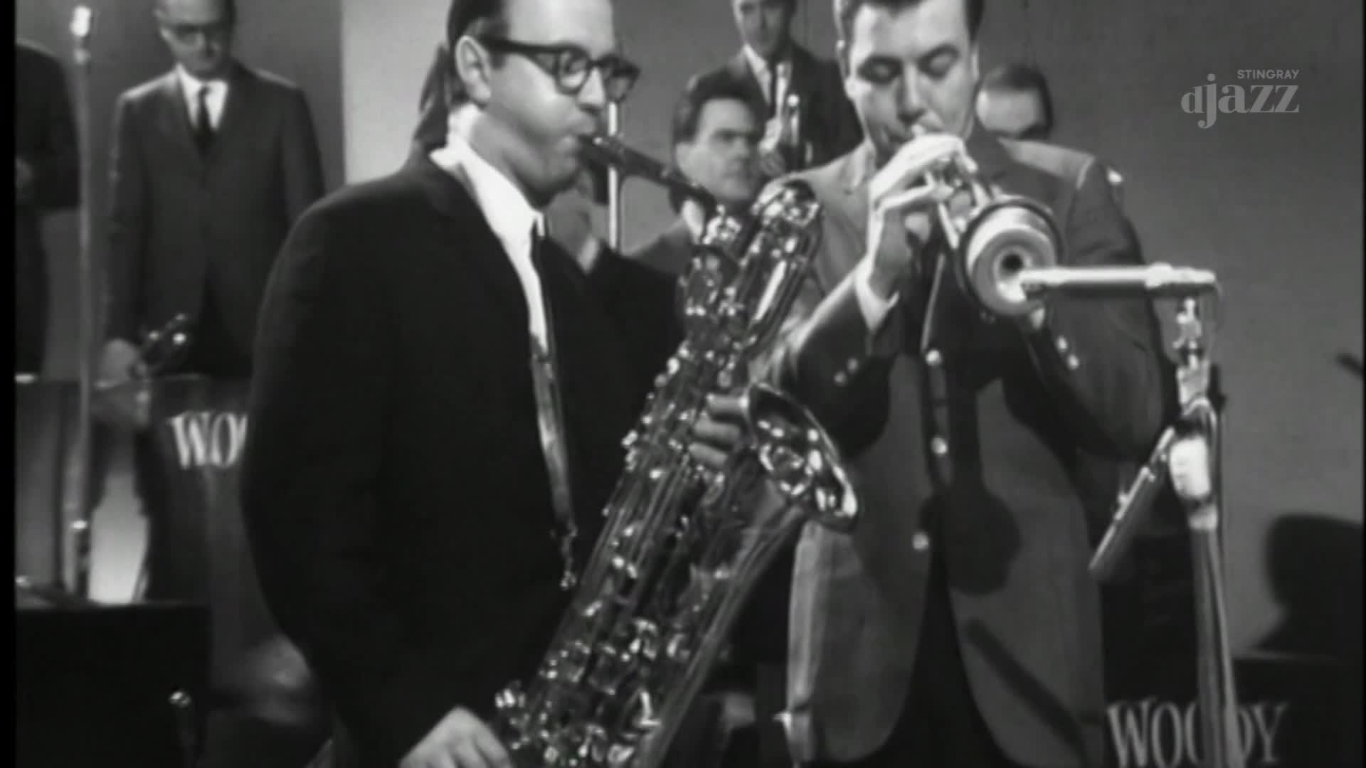 A man in a suit plays a tenor saxophone, his fingers moving over the keys. Beside him, another musician blows into a trumpet, the bell pointed towards a microphone.