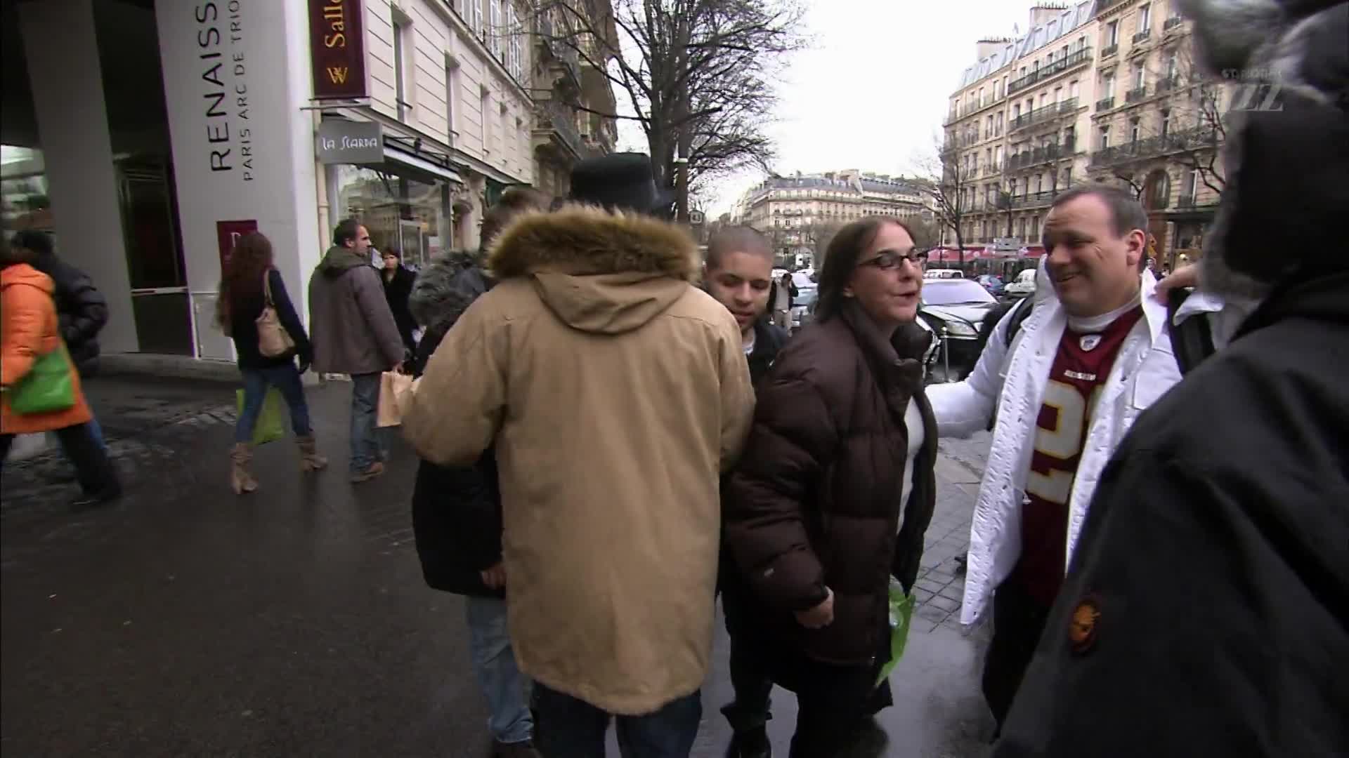 A woman in a brown puffer coat talks animatedly with a man wearing a white jacket over a burgundy jersey. They stand on a wet sidewalk beside a building with "RENAISSANCE PARIS ARC DE TRIOMPHE" printed on its facade.