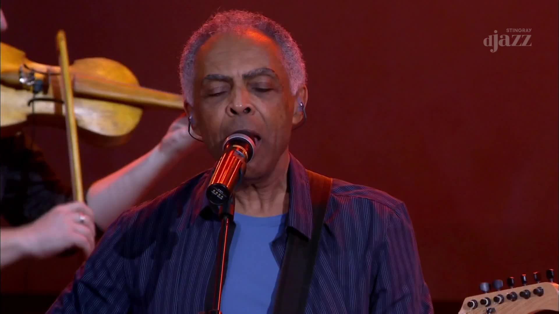 The singer's eyes are closed as he sings into the microphone, his guitar slung across his chest. A violinist plays in the background, their bow moving across the strings.