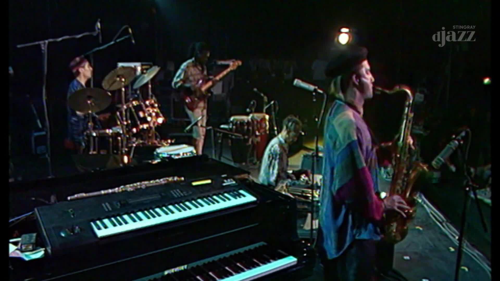 A saxophonist in a colorful shirt plays his instrument while a guitarist stands nearby. The drummer is seated behind a kit, and a keyboardist works at his console.