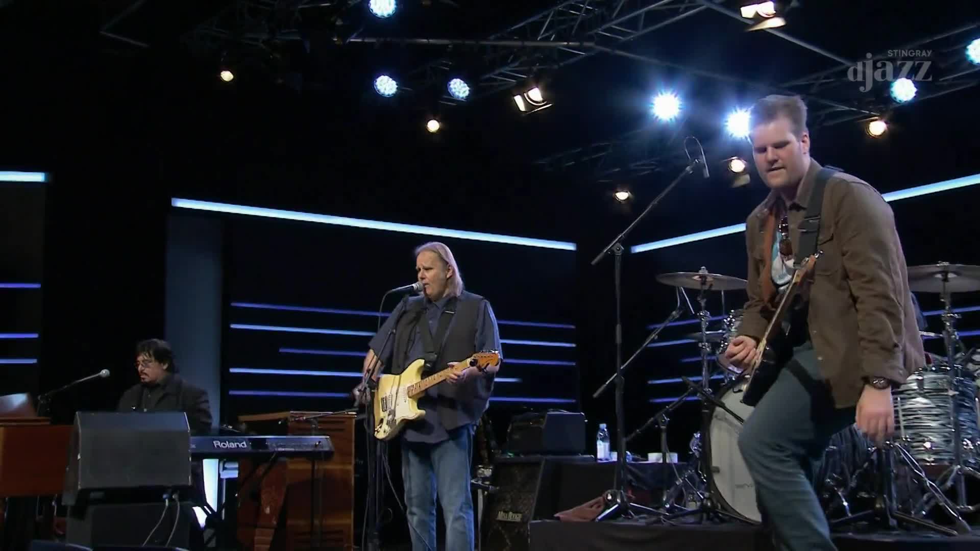 A guitarist in a brown jacket steps forward, his instrument angled towards the audience. To his left, a singer strums a light-colored electric guitar, his voice amplified by a nearby microphone.