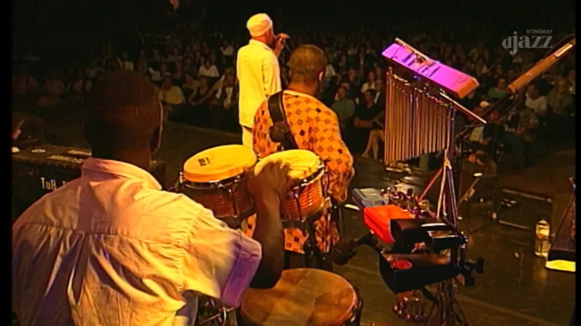 A drummer plays bongo drums, his hands a blur of motion. Upstage, a singer in white addresses the audience as a vibraphone stands ready.