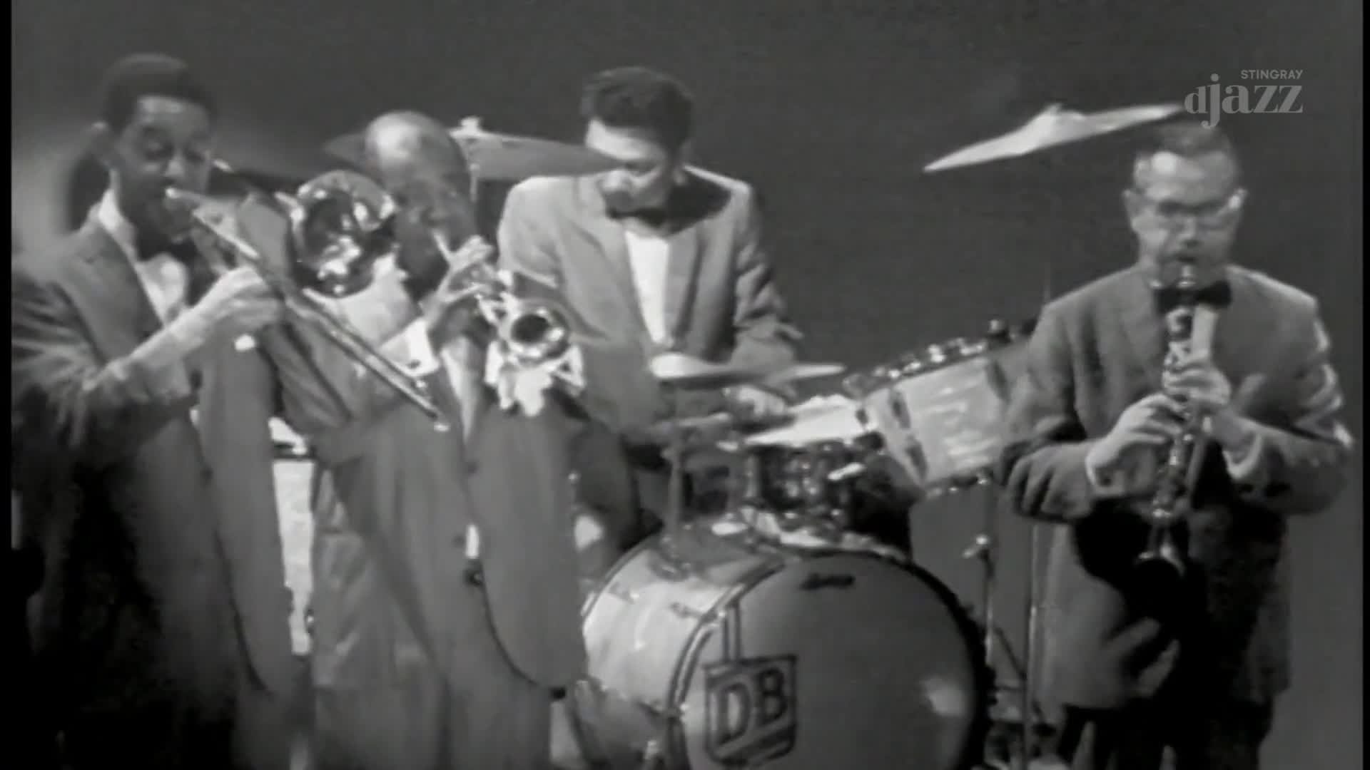 A trombonist in a sharp suit leans into his instrument, while a drummer in a similar suit keeps time. To the right, a clarinetist with glasses plays his part.