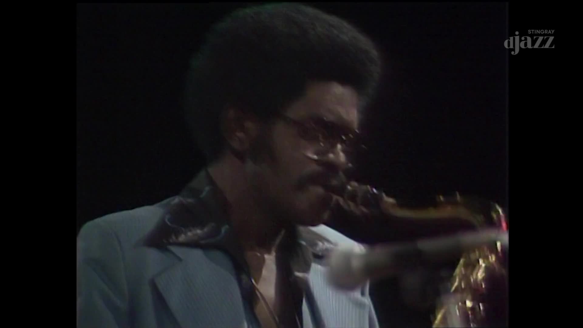 A man with a large afro and sunglasses plays a saxophone, his light blue jacket catching the stage lights. The Stingray DJAZZ logo appears in the upper right corner.