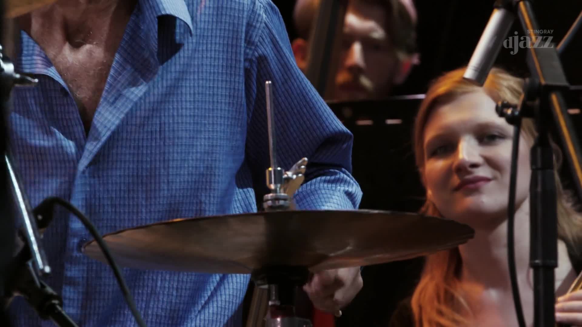 A drummer in a blue checkered shirt prepares to strike a cymbal, his hand poised. Behind him, a young woman with long auburn hair smiles, her violin resting on her shoulder.
