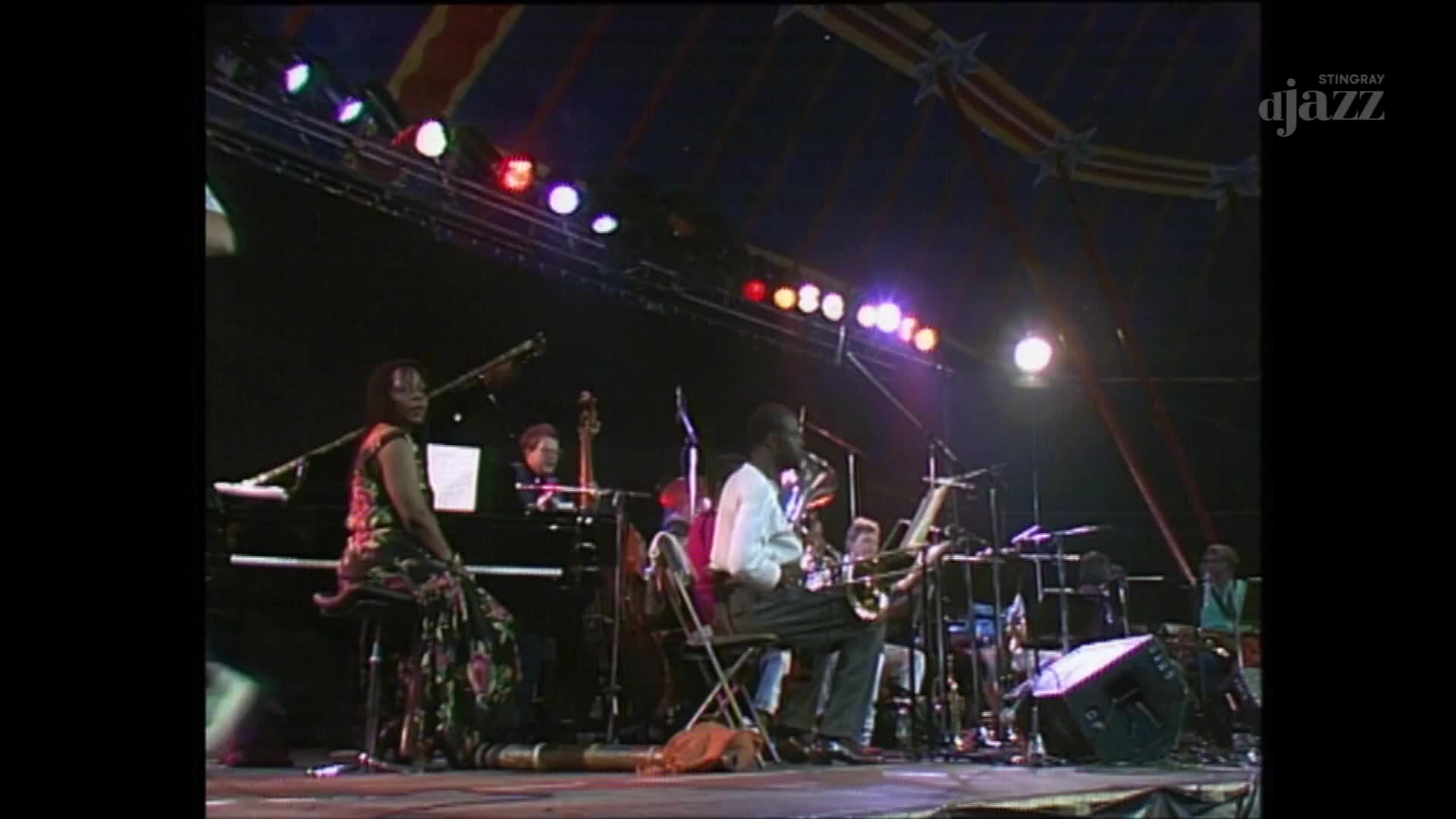 A jazz quartet is performing under a tent at a Canadian festival. The saxophonist, in a white shirt, plays a solo while the pianist, in a vibrant dress, watches him.