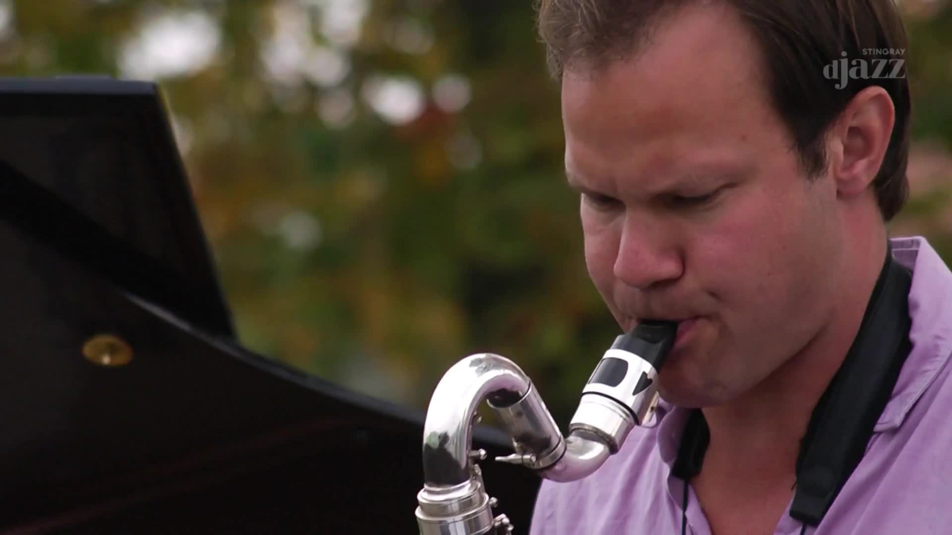 A man in a lavender shirt plays a saxophone outdoors, the instrument's silver curves catching the light. The Stingray DJAZZ logo is visible in the background, suggesting a Canadian broadcast.