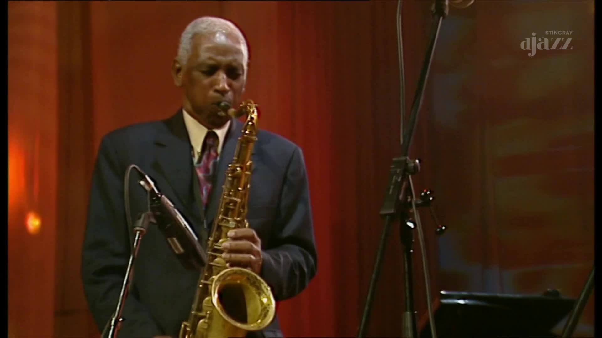 A distinguished gentleman in a suit plays a golden saxophone, his fingers moving nimbly over the keys. The warm, red backdrop suggests a performance for Stingray DJAZZ, perhaps here in Canada.