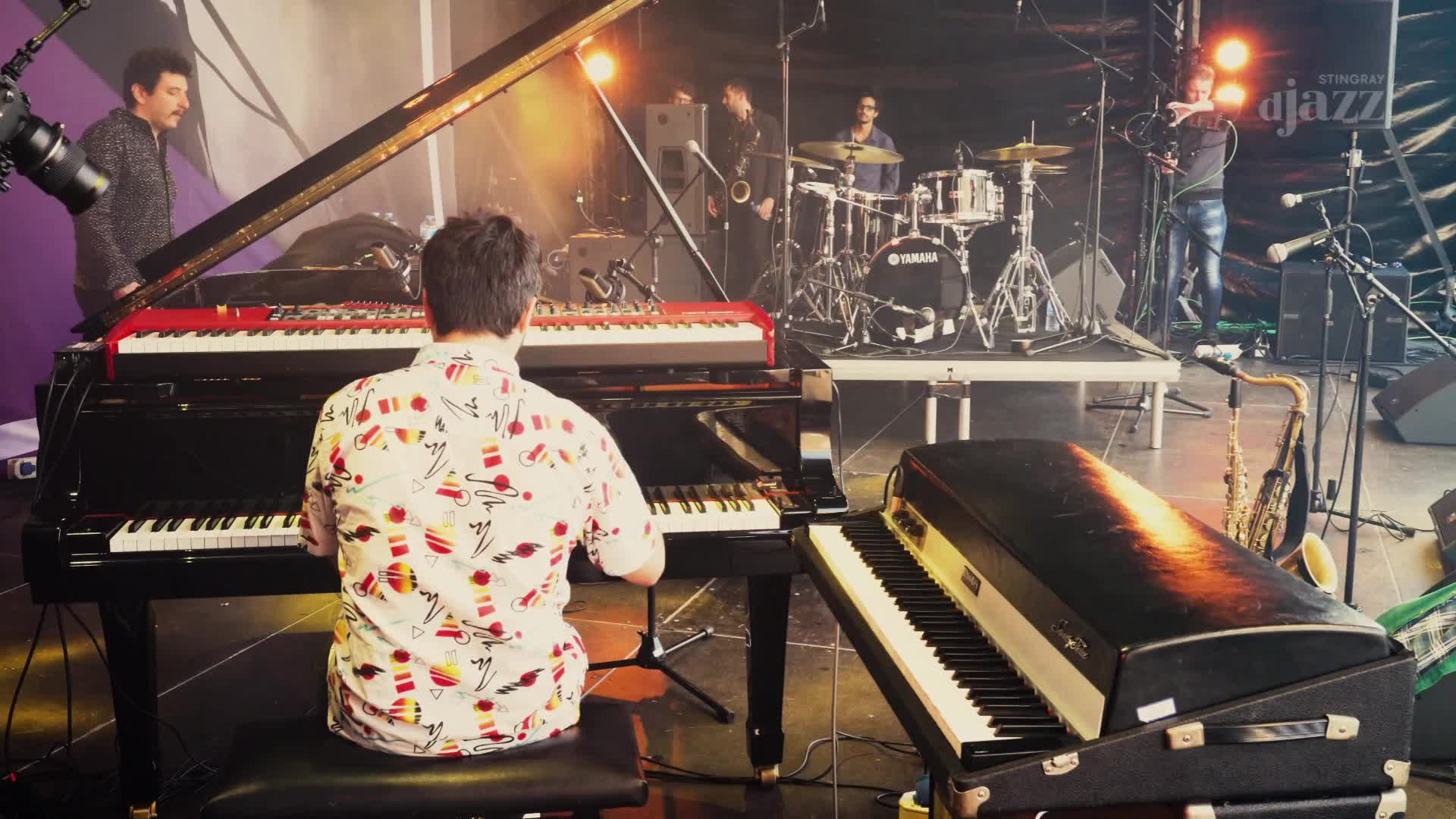 A pianist in a colourful shirt plays a grand piano, while a saxophonist and drummer perform behind him on a stage set up for Stingray DJAZZ. The band is playing outdoors, likely in Canada, with a bright spotlight illuminating the scene.