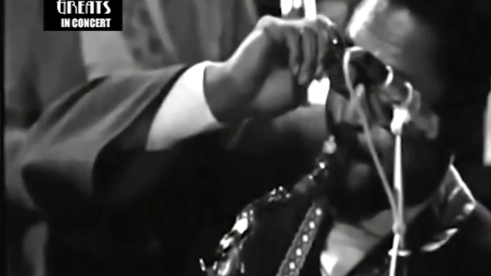 A man in sunglasses leans into his saxophone, his fingers poised over the keys. The "Greats in Concert" title card suggests a Canadian broadcast.