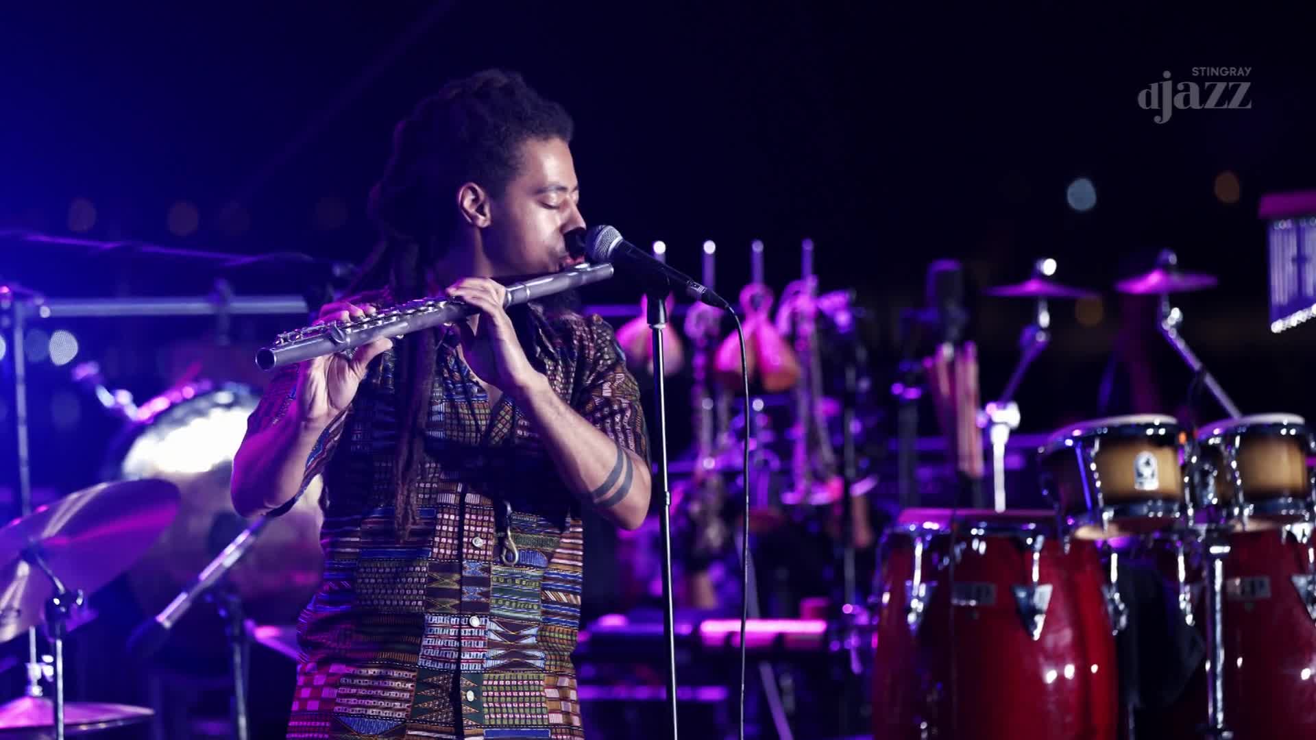 A musician plays a silver flute, his fingers moving across the keys. Behind him, a collection of conga drums and other percussion instruments gleam under the stage lights. A musician plays a silver flute, his fingers moving across the keys. Behind him, a collection of conga drums and other percussion instruments gleam under the stage lights.
