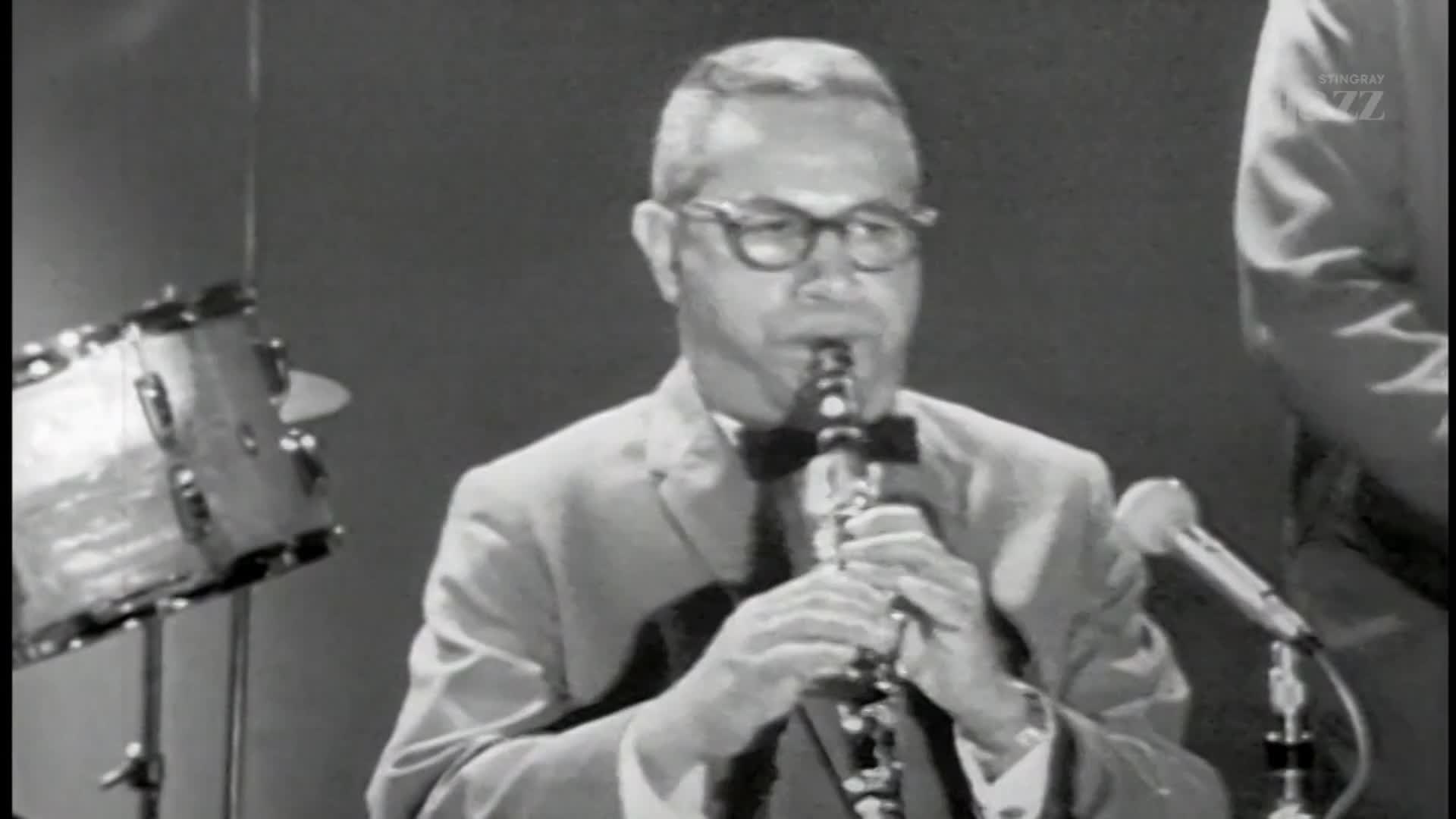A man in a suit and bowtie plays a clarinet, his fingers moving nimbly over the keys. To his left, a drummer's snare sits ready. A man in a suit and bowtie plays a clarinet, his fingers moving nimbly over the keys. To his left, a drummer's snare sits ready.