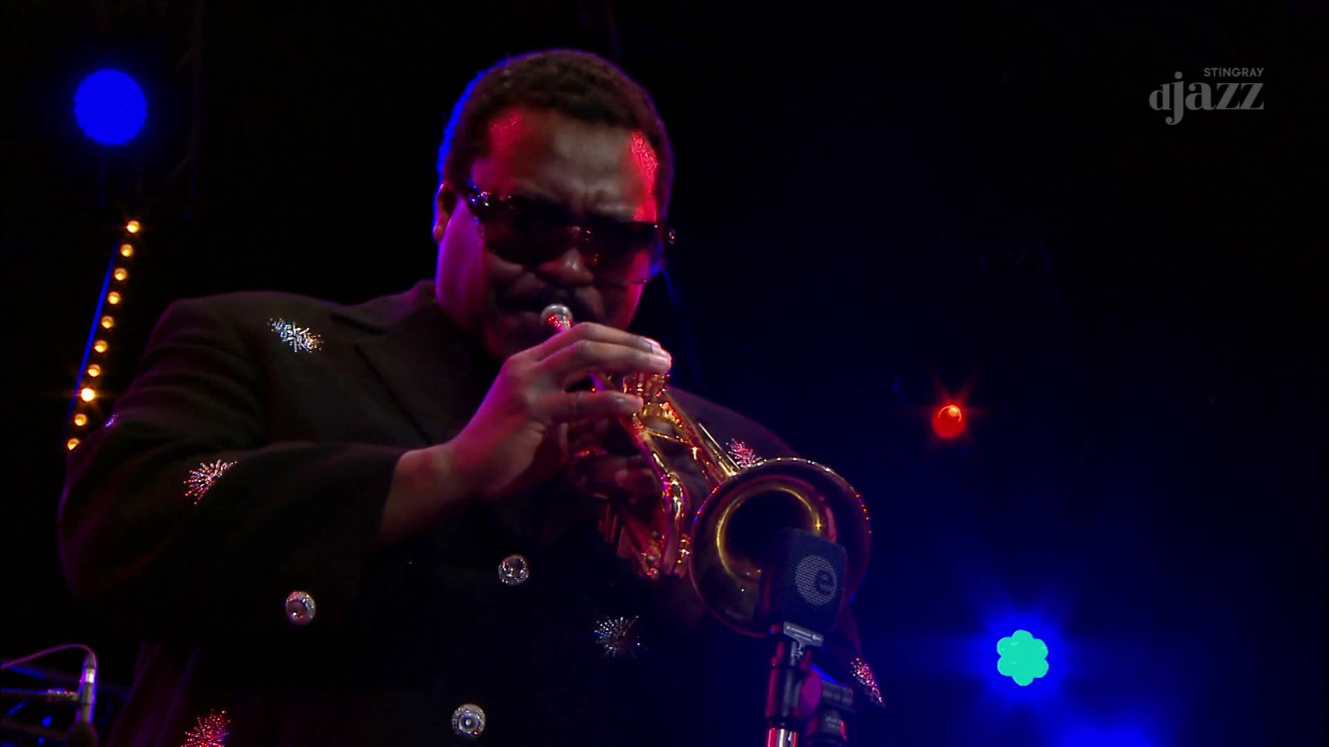 A trumpeter in a sparkling black jacket plays his instrument under the stage lights. The Stingray Djazz logo appears in the upper right corner. A trumpeter in a sparkling black jacket plays his instrument under the stage lights. The Stingray Djazz logo appears in the upper right corner.